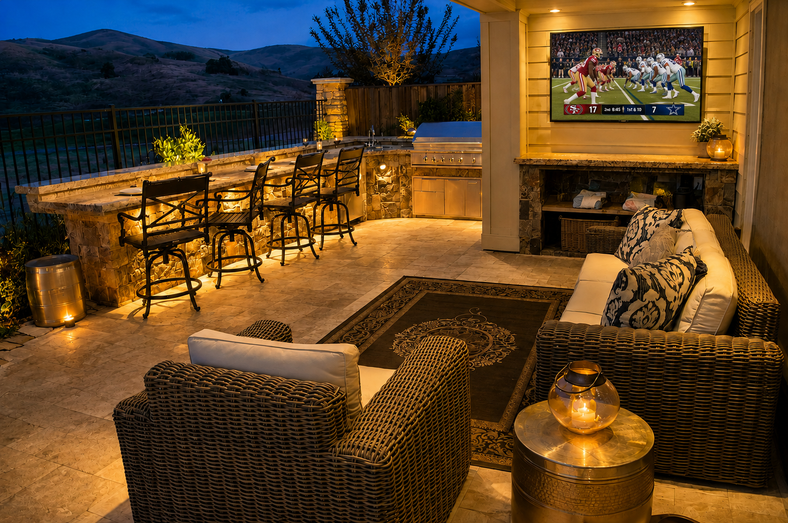 A cozy outdoor patio at dusk with a mountain view. The space features a bar counter with seating, a fireplace, a TV showing a football game, and comfortable wicker chairs with cushions, illuminated by candles and warm lighting.