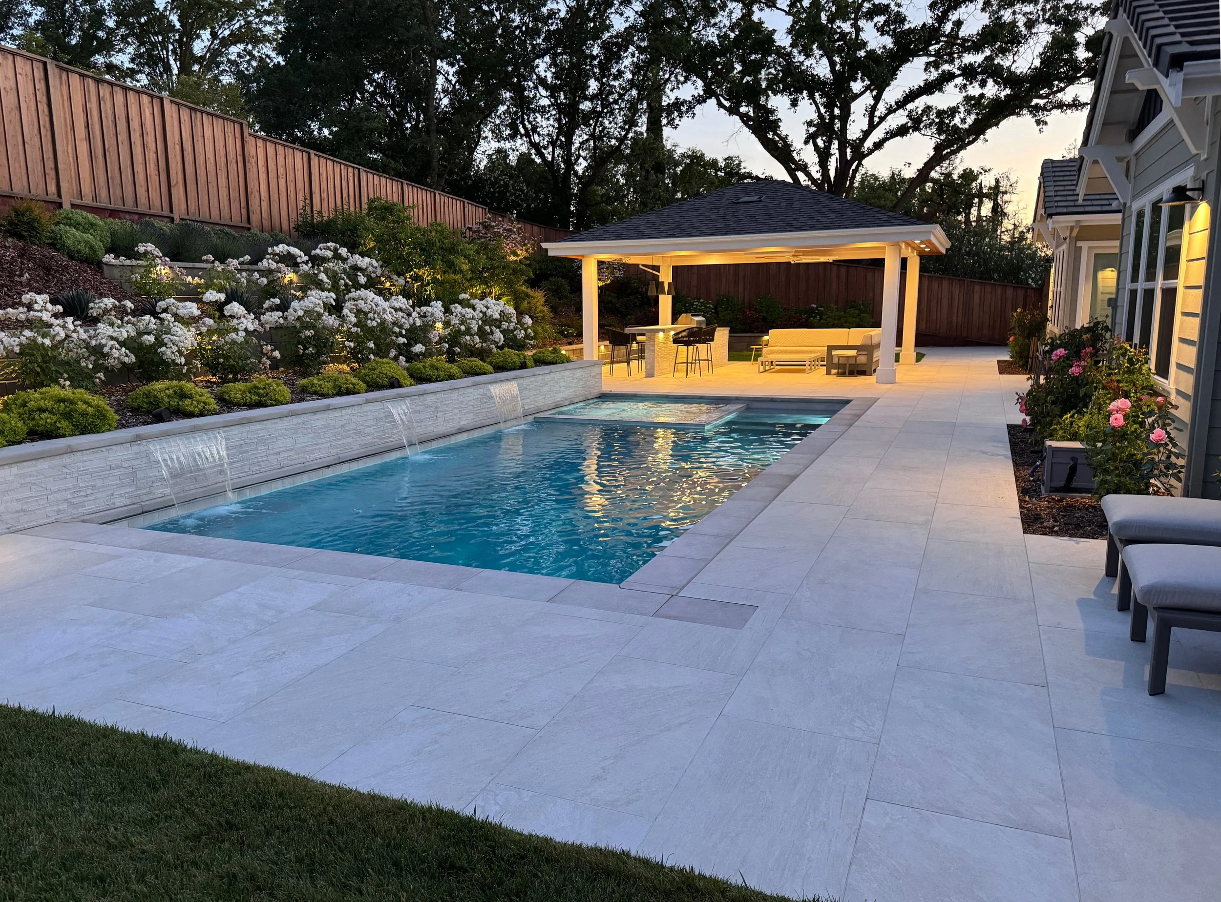 A backyard with a swimming pool, outdoor seating area, garden with white and pink flowers, wooden fence, and large trees at dusk.
