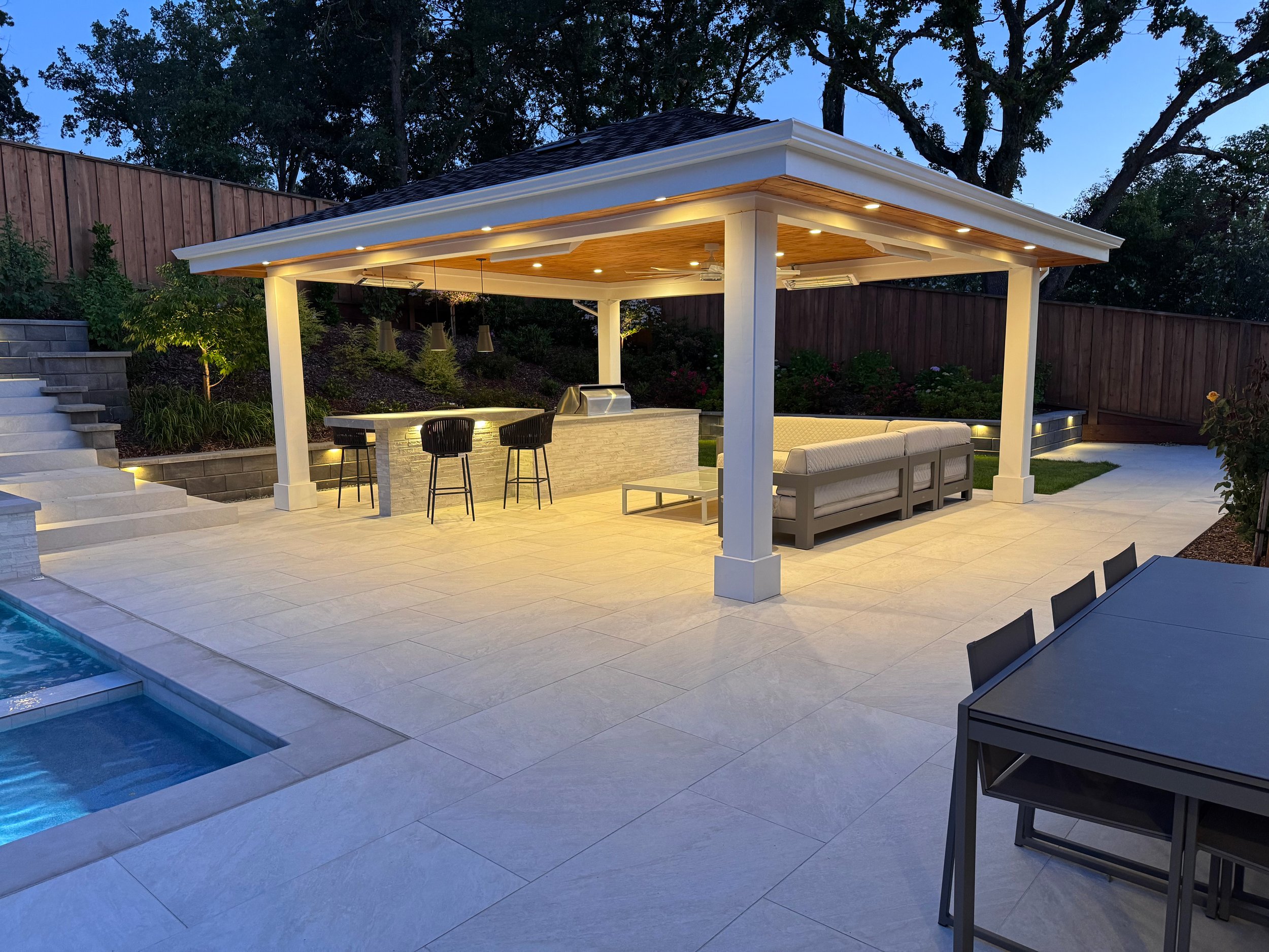 A modern outdoor patio area with a pergola, bar seating, sofa, and outdoor lighting, next to a swimming pool at dusk.