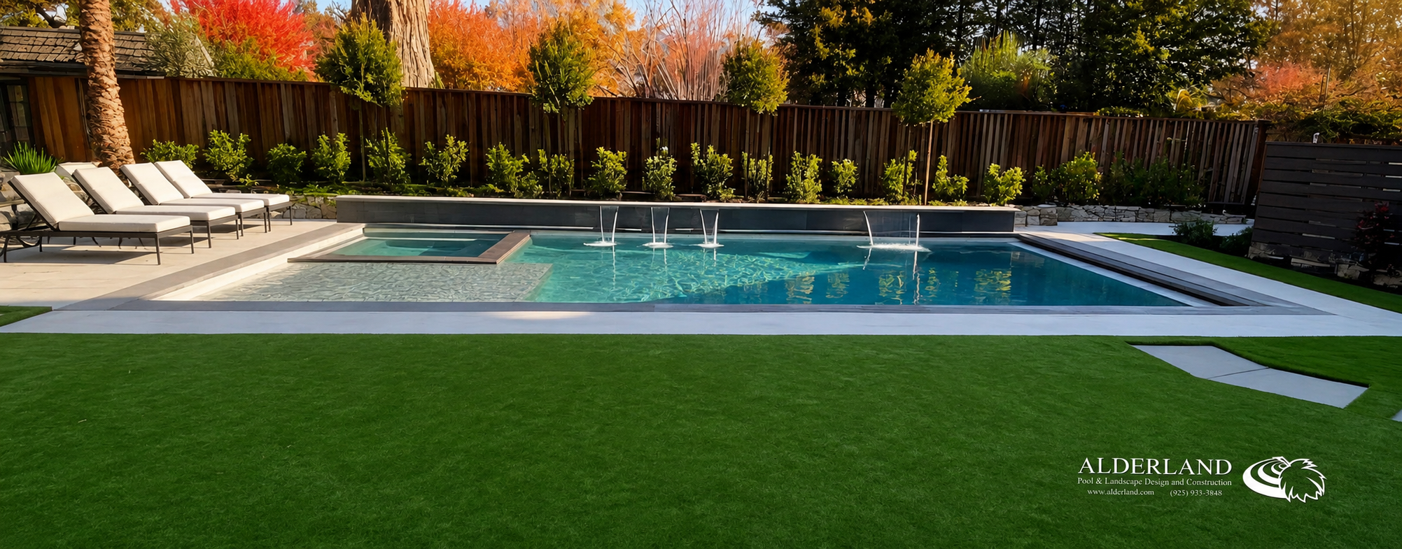 Backyard with a swimming pool featuring a built-in hot tub, surrounded by a green lawn, lounge chairs, and landscaped bushes and trees with fall foliage.