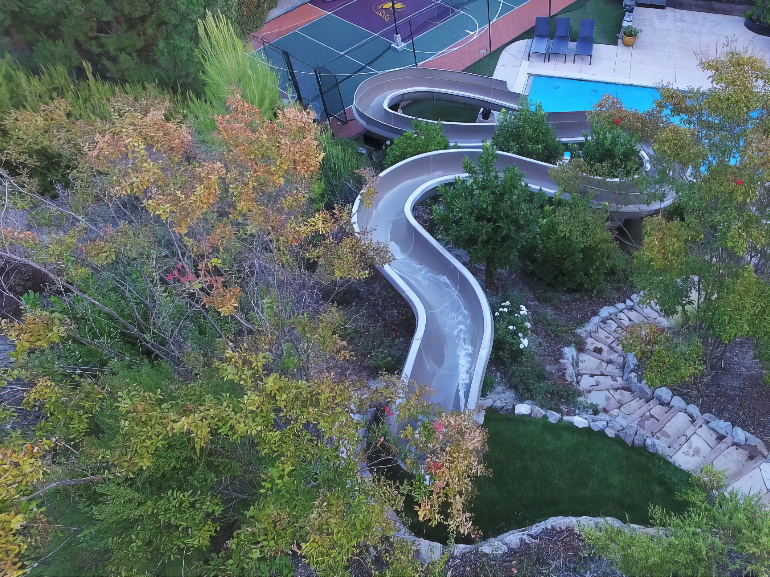 A tree-lined playground with a large waterslide, swim pool, tennis court, and seating area.