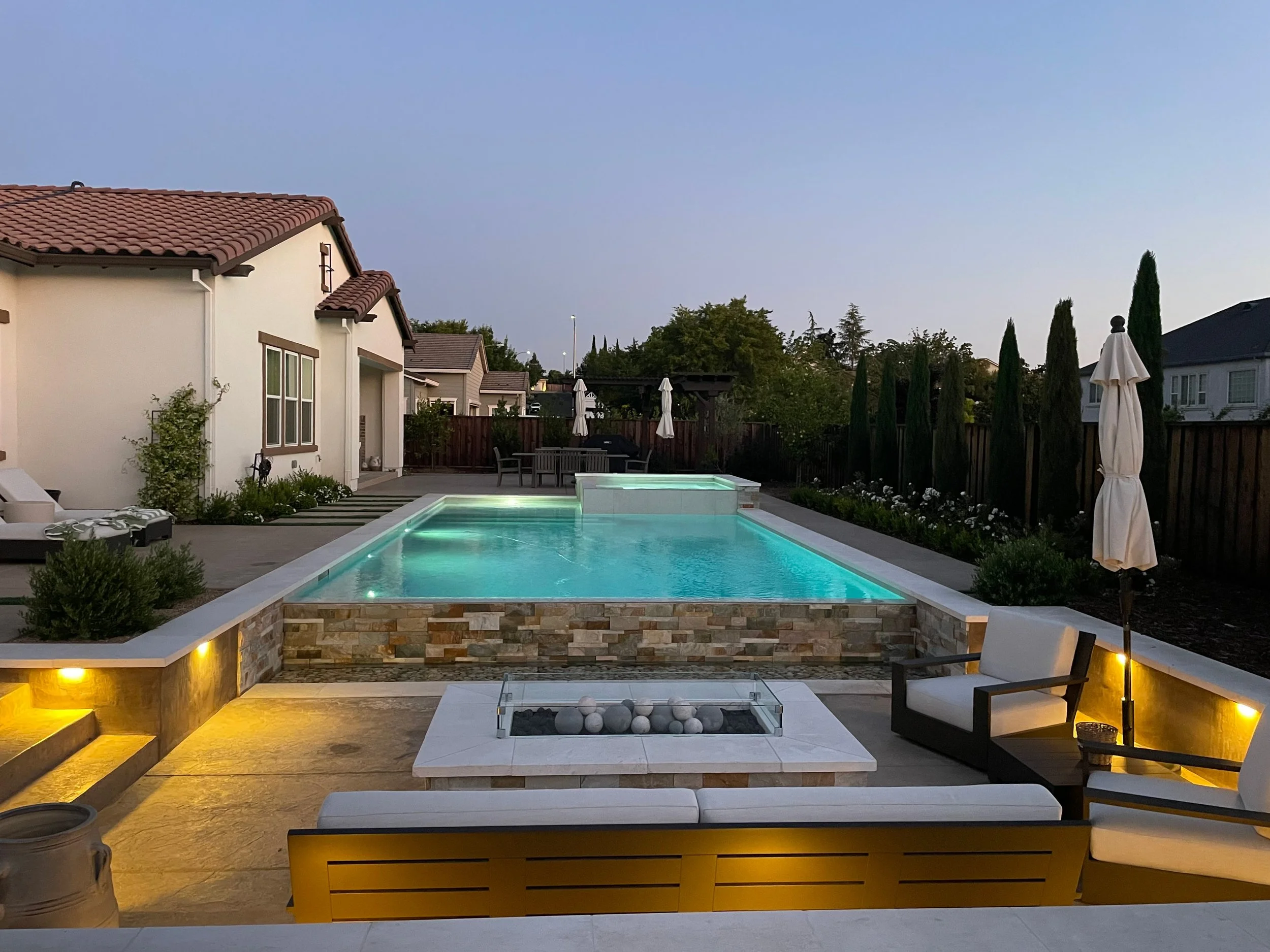 Resort-style backyard with a lit swimming pool, surrounded by patio furniture, umbrellas, plants, and trees at dusk.
