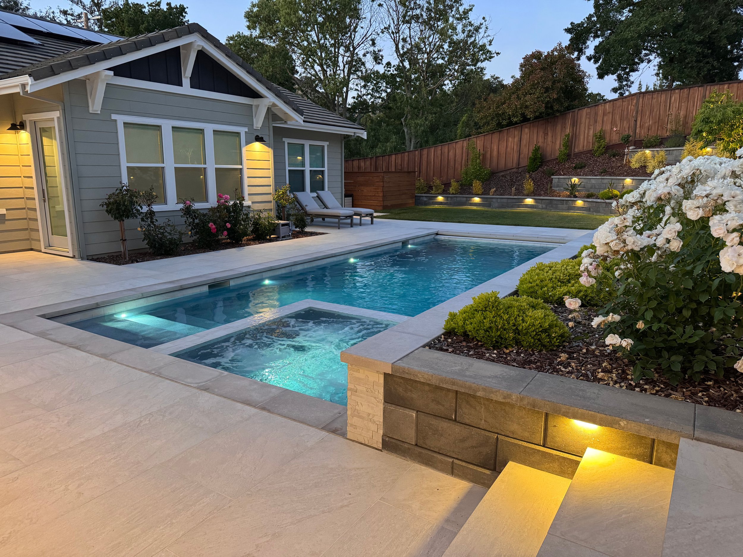 A backyard with a modern house, outdoor pool with built-in lighting, lounge chairs, landscaped garden beds with white flowers and shrubs, terraced yard with lighting, and a wooden fence, all illuminated during dusk.