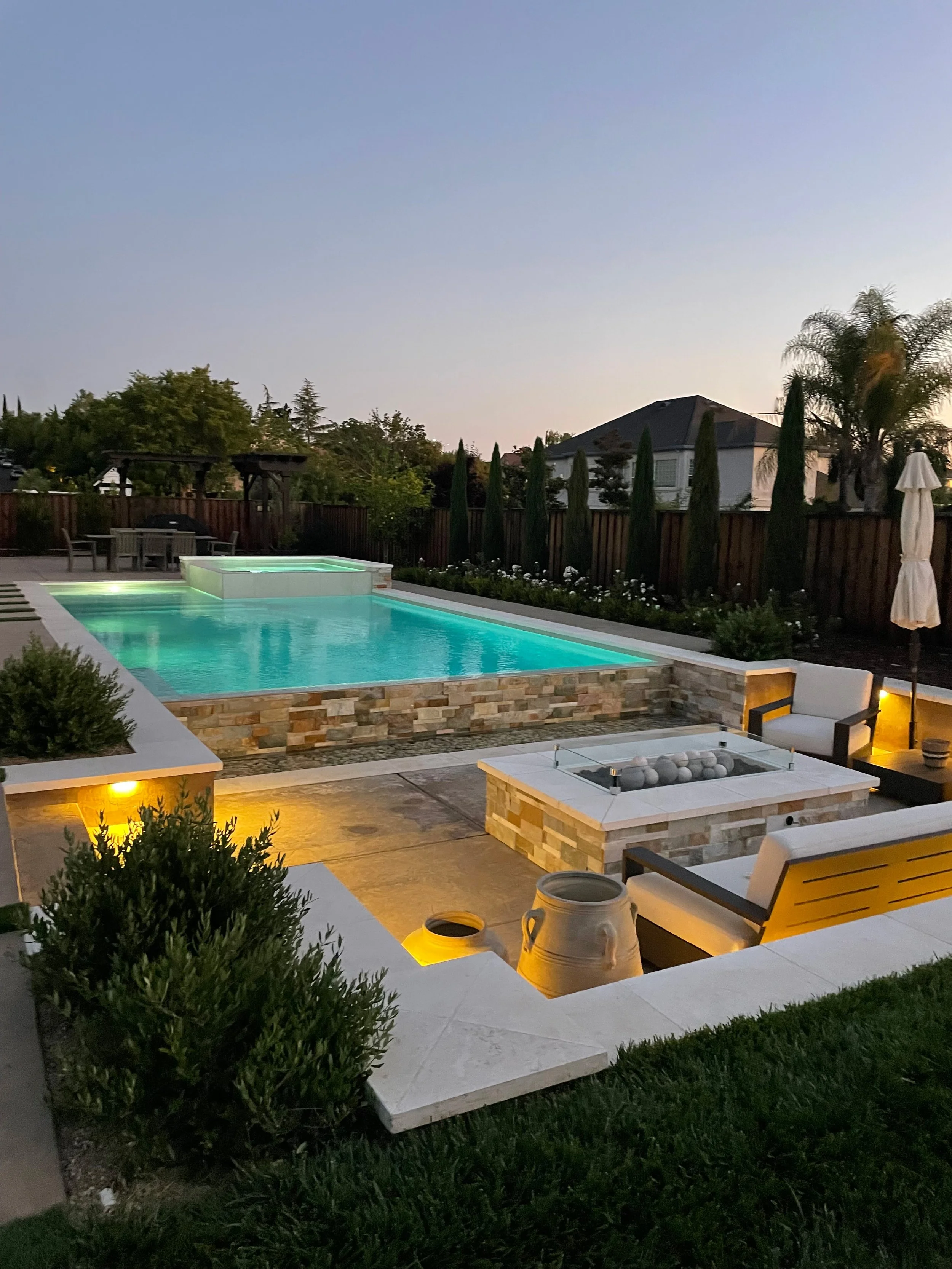 A backyard swimming pool area at dusk, with lounge chairs, a fire pit, a covered umbrella, and landscaped plants.