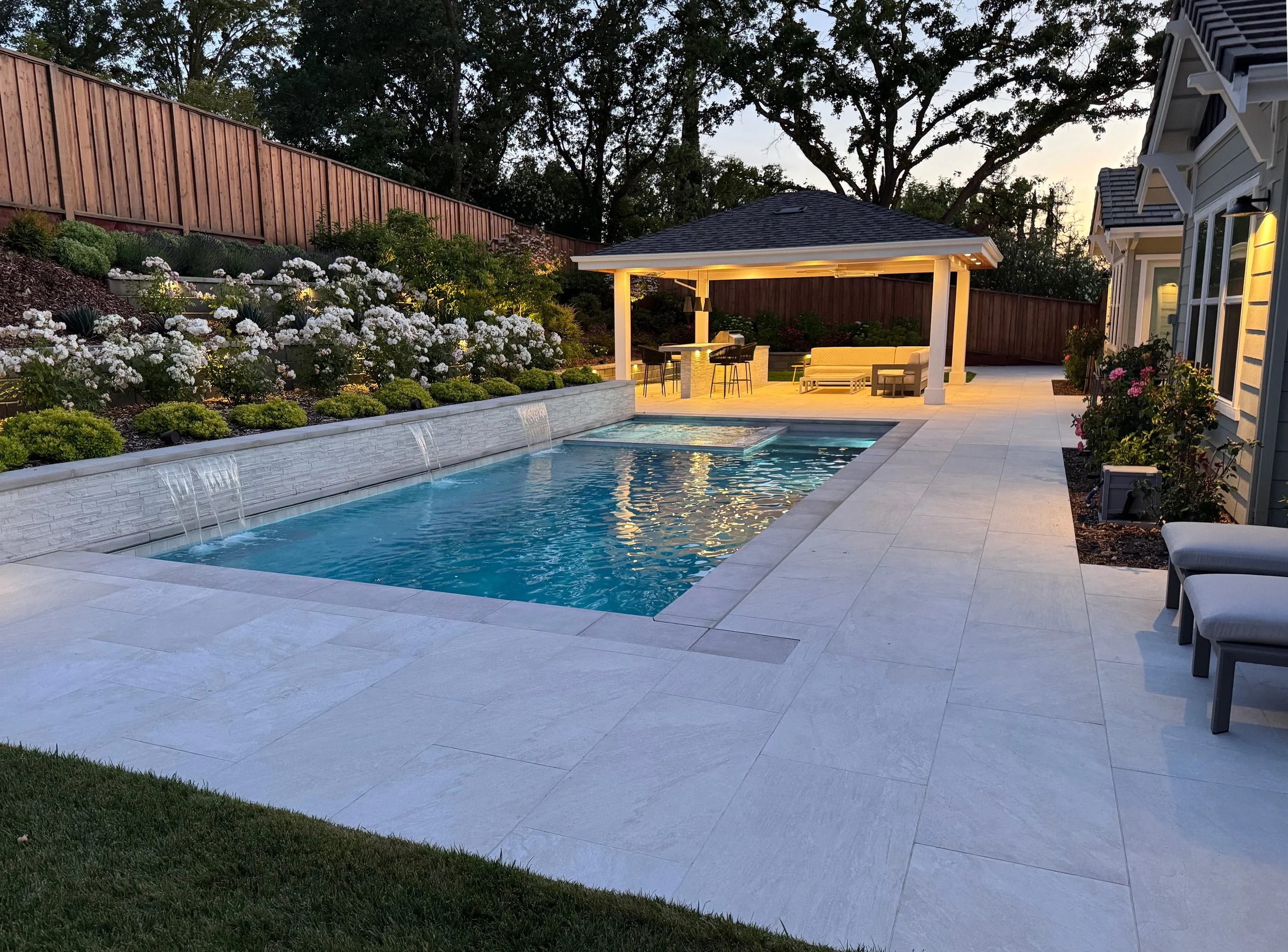 A backyard with a lit swimming pool, a pavilion with outdoor furniture, flowering shrubs, and a wooden fence in the evening.