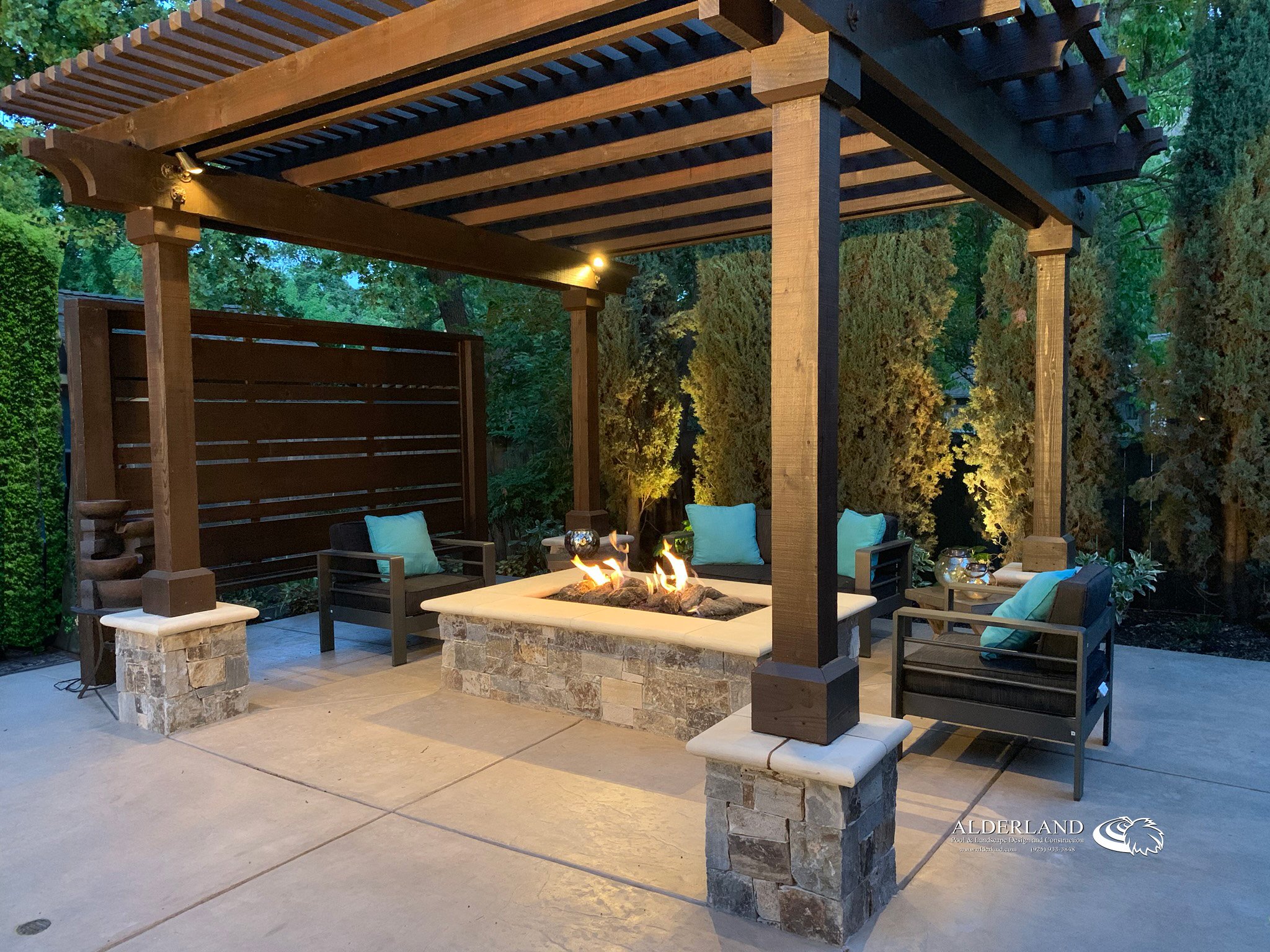 Outdoor patio area with a fire pit, seating, and a wooden pergola surrounded by trees and greenery.