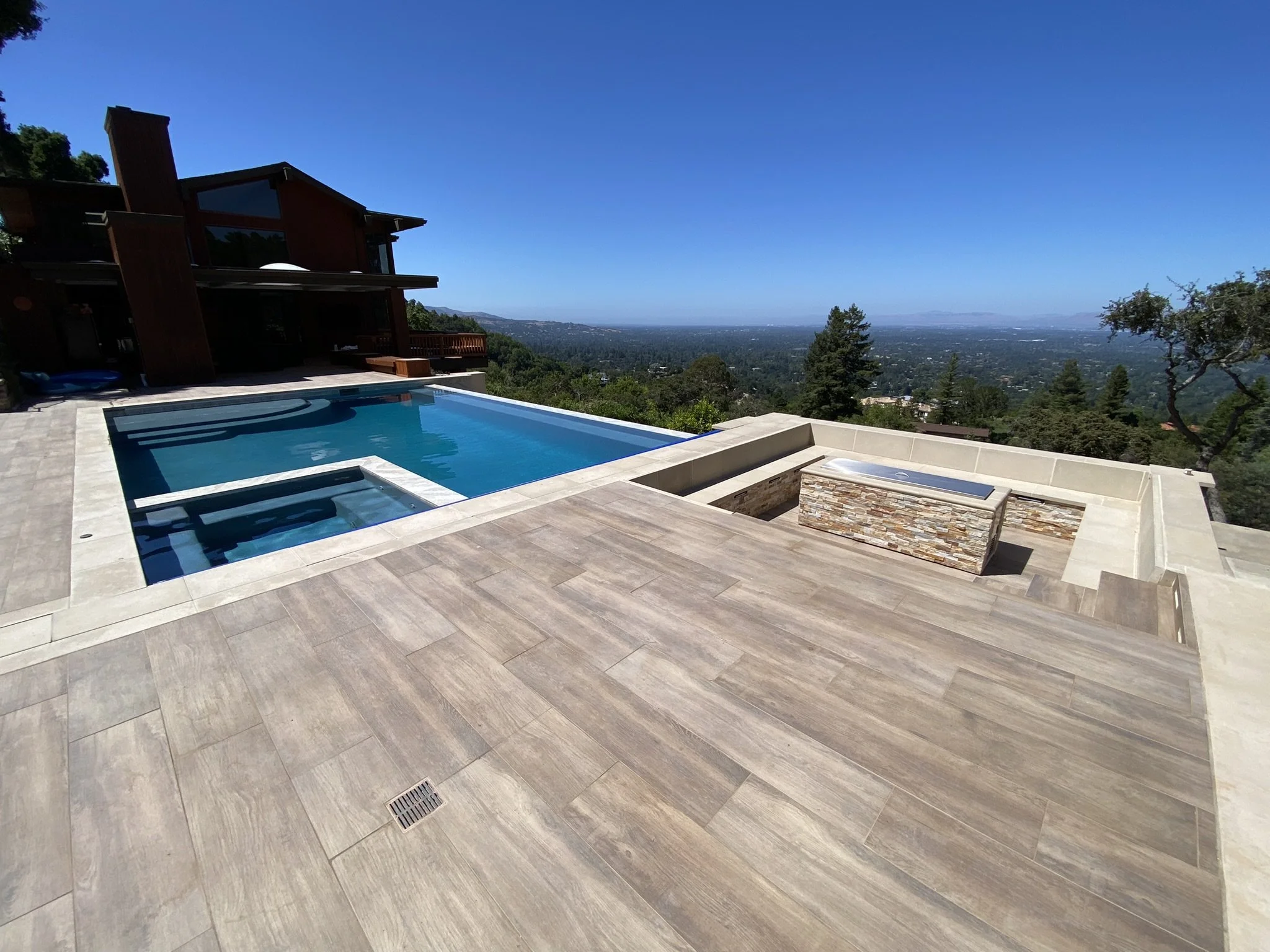 Modern outdoor pool area with view over a city and forest, featuring a rectangular swimming pool, a hot tub, and a hot tub cover, surrounded by wooden and stone flooring, with a contemporary house in the background.