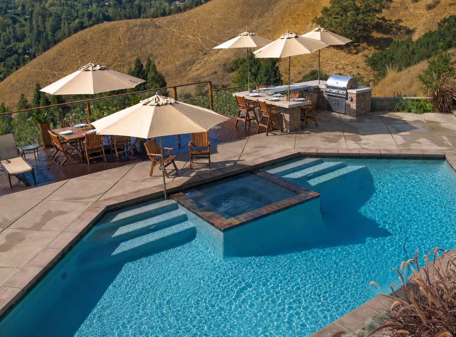 Poolside outdoor dining area with umbrellas, chairs, and a grill, overlooking dry hills and trees in the background.