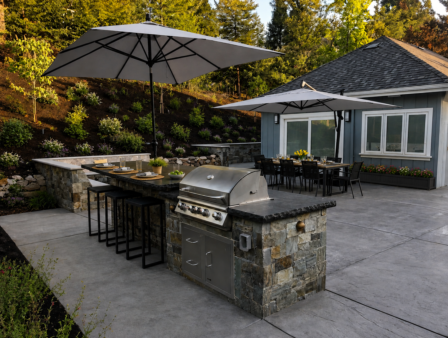 An outdoor patio with a stone barbecue grill, a dining table with chairs, and large umbrellas, surrounded by gardens and a house with blue siding.