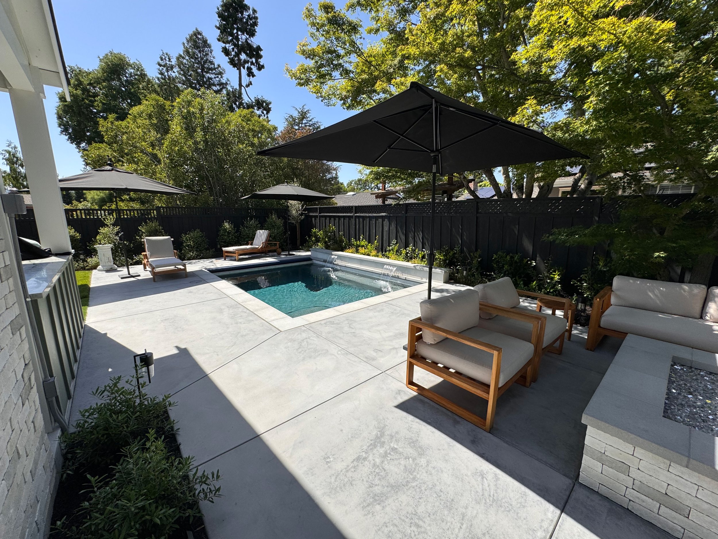 Backyard with a small swimming pool, patio furniture with cushions and umbrellas, surrounded by green trees and a black privacy fence.
