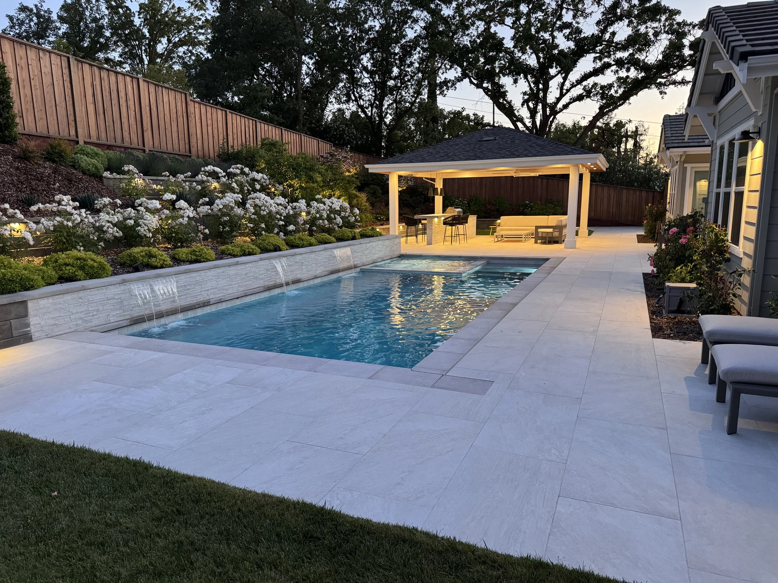 Backyard with swimming pool, landscaped garden with white flowers, outdoor seating area under a gazebo, neighboring house with exterior lights, and a wooden privacy fence.