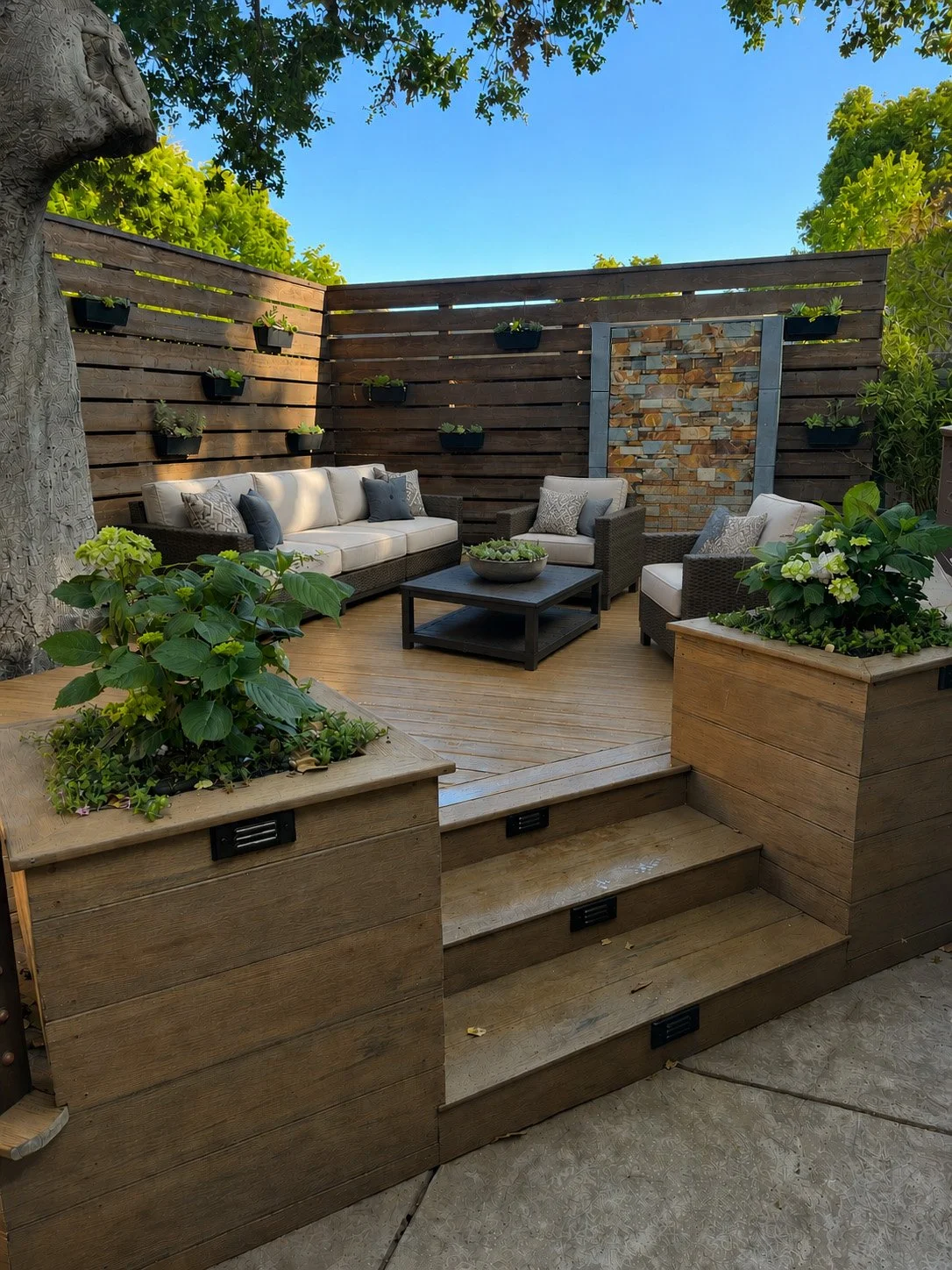 A cozy outdoor patio with wooden flooring, outdoor sofa, armchairs, coffee table, and lush potted plants against a wooden privacy fence with decorative wall feature and hanging planters, under clear blue sky.