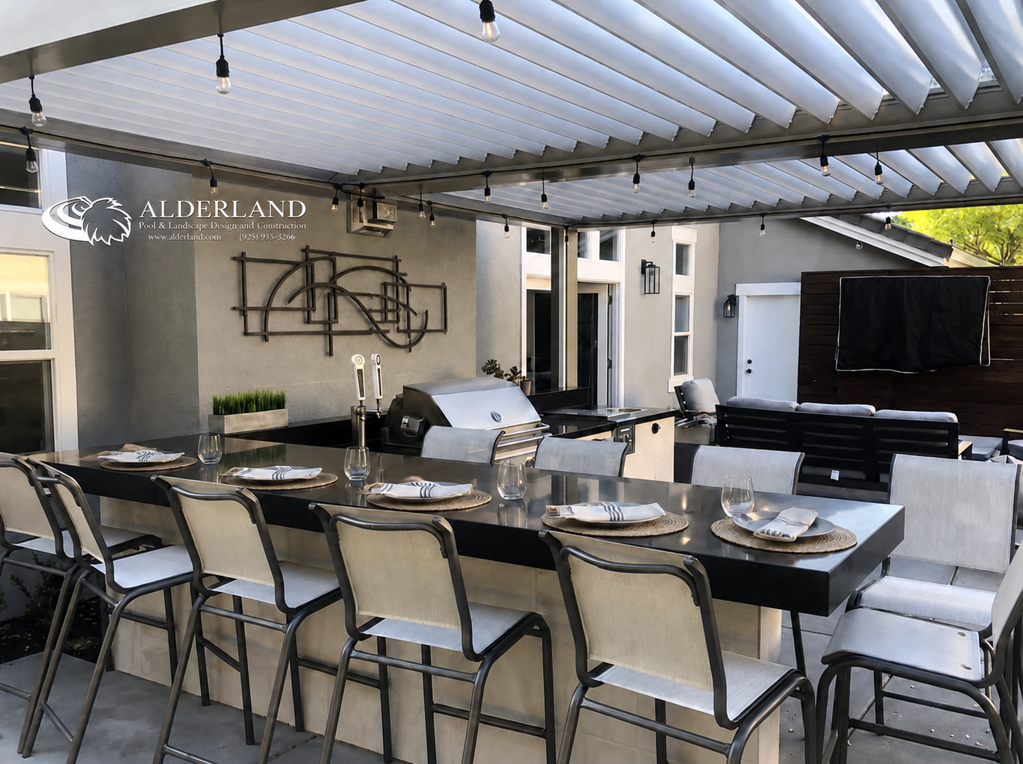 Outdoor patio with a long dining table set with plates, napkins, utensils, and glasses, under a partially covered roof with string lights. There's a built-in grill, wall art, and seating area with outdoor furniture.