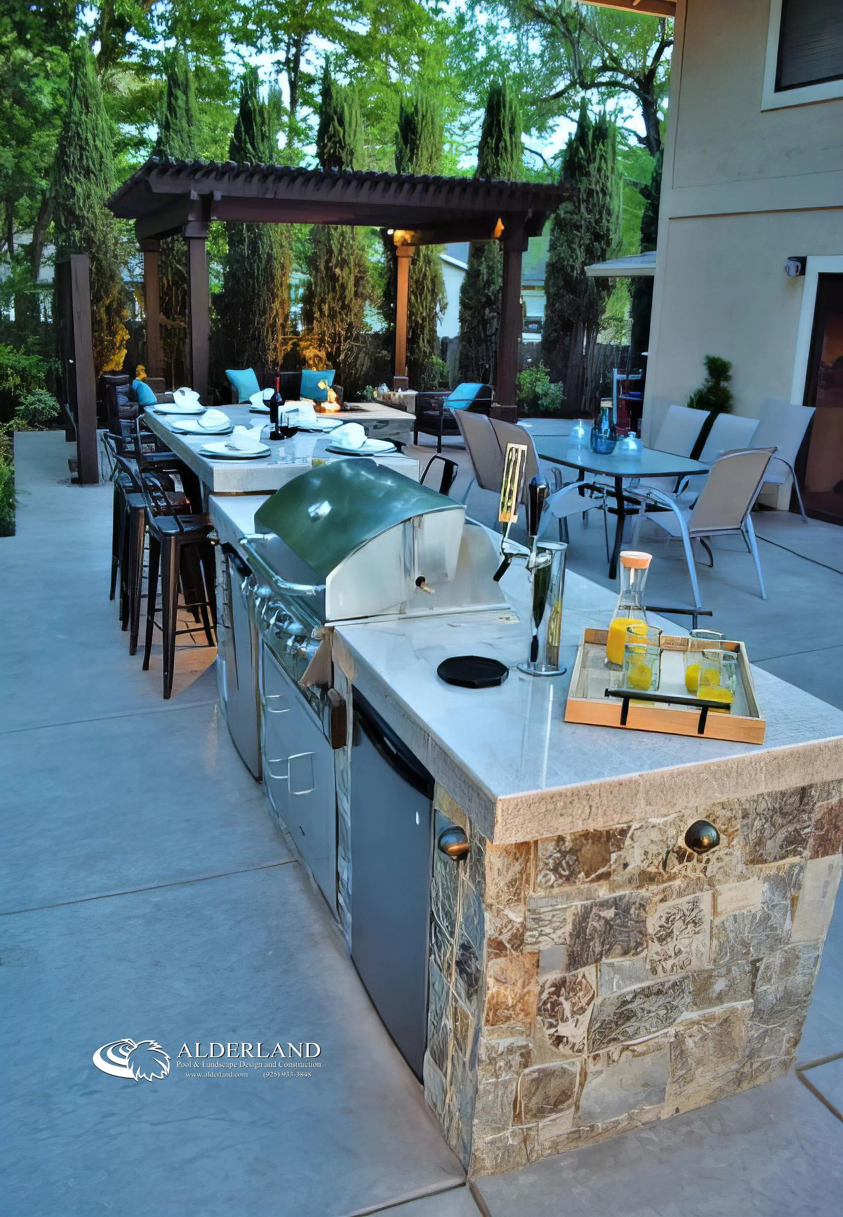 Outdoor kitchen with a granite counter, stainless steel appliances, and a stone base, set on a patio with a dining area under a wooden pergola surrounded by trees.