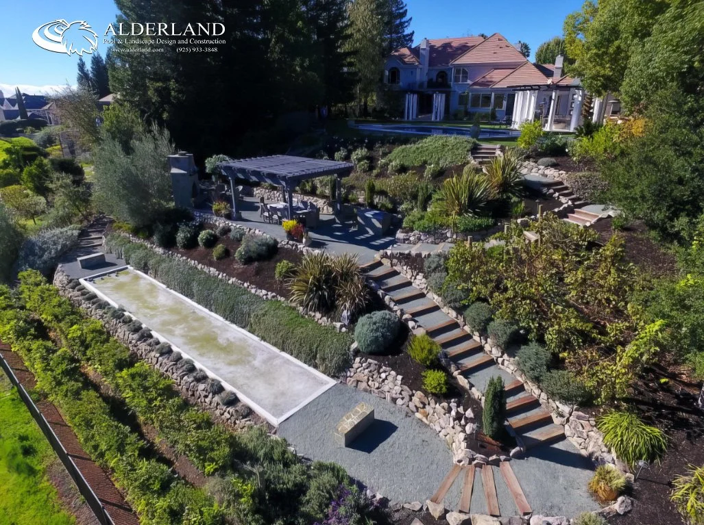 A landscaped backyard with a tiered garden, stone steps, and a covered patio area in front of a large house. The yard features various plants, shrubs, and a long rectangular water feature.