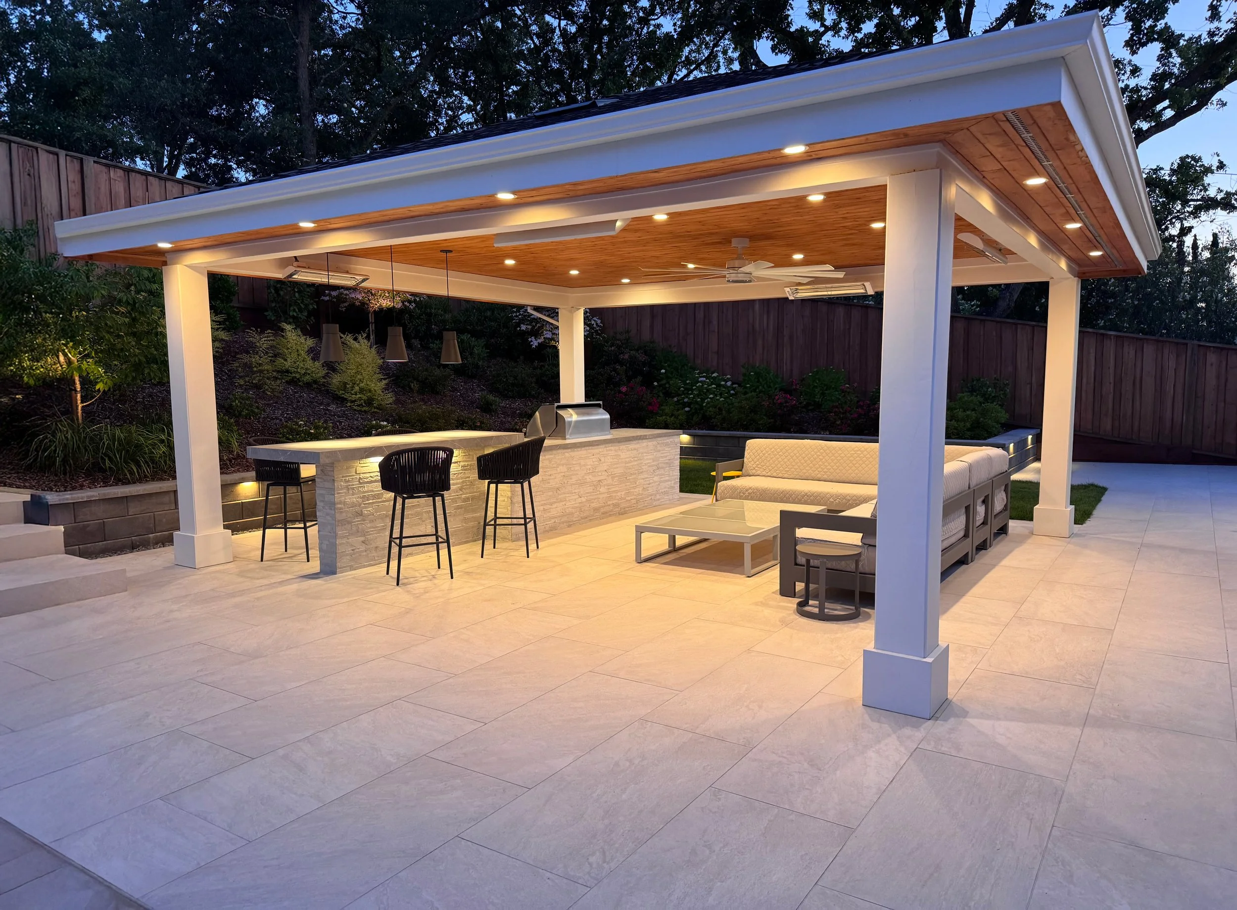 Outdoor patio with a covered roof, built-in bar area with three black chairs, a sofa, a coffee table, and a small round side table, surrounded by greenery and a wooden fence.