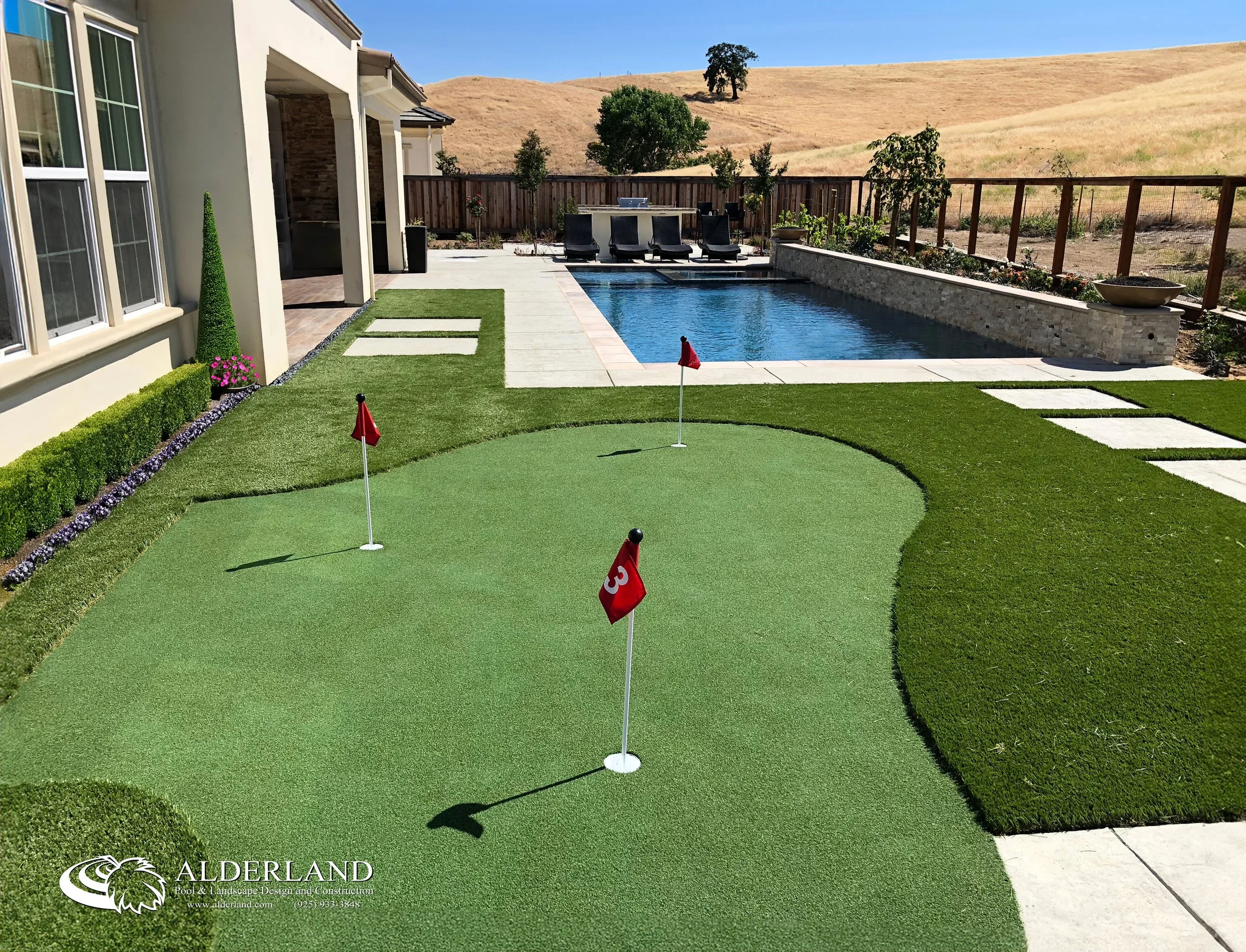 A backyard with an artificial putting green, flags, and a pool with a stone edge. There are lounge chairs near the pool, a garden bed with plants, and a fenced yard with rolling hills in the background.