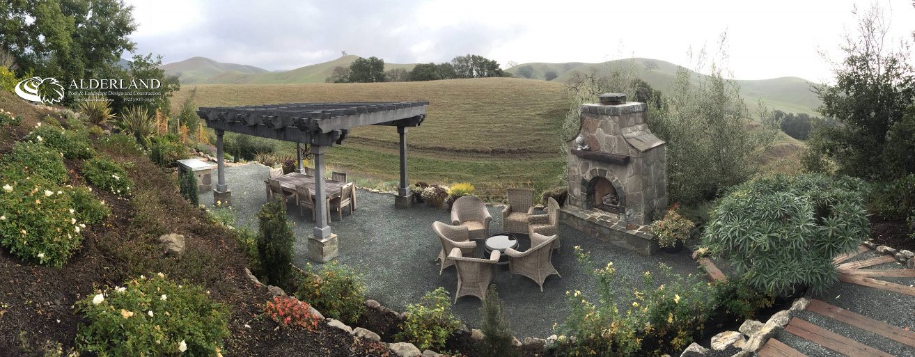 Outdoor patio with seating area, stone fireplace, pergola, surrounded by plants, with hills in the background.