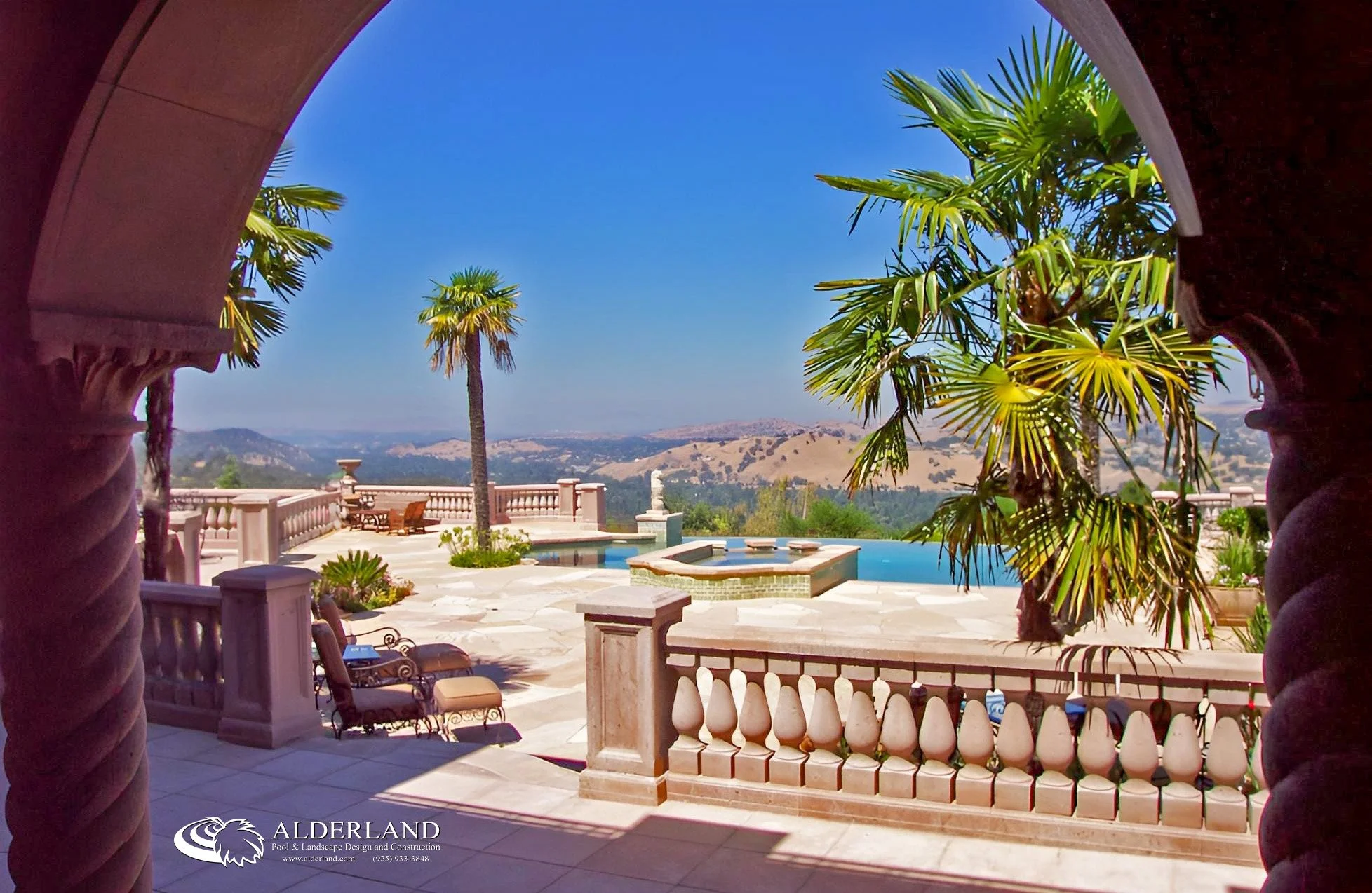 A spacious balcony with outdoor furniture and a hot tub, overlooking rolling hills and a clear blue sky. Palm trees and decorative plants add to the luxurious landscape view.