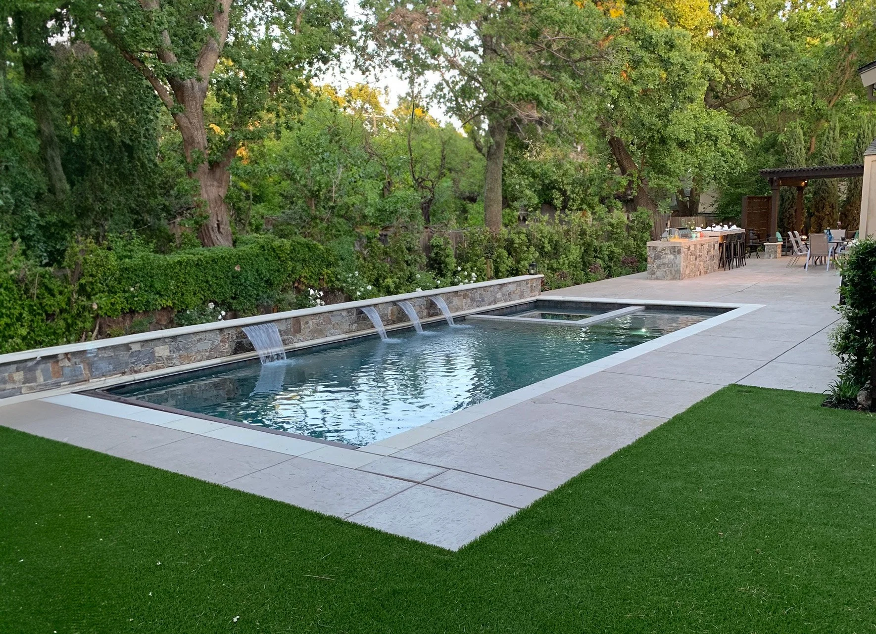 Backyard with in-ground swimming pool, lush green trees, and outdoor dining area with table and chairs.