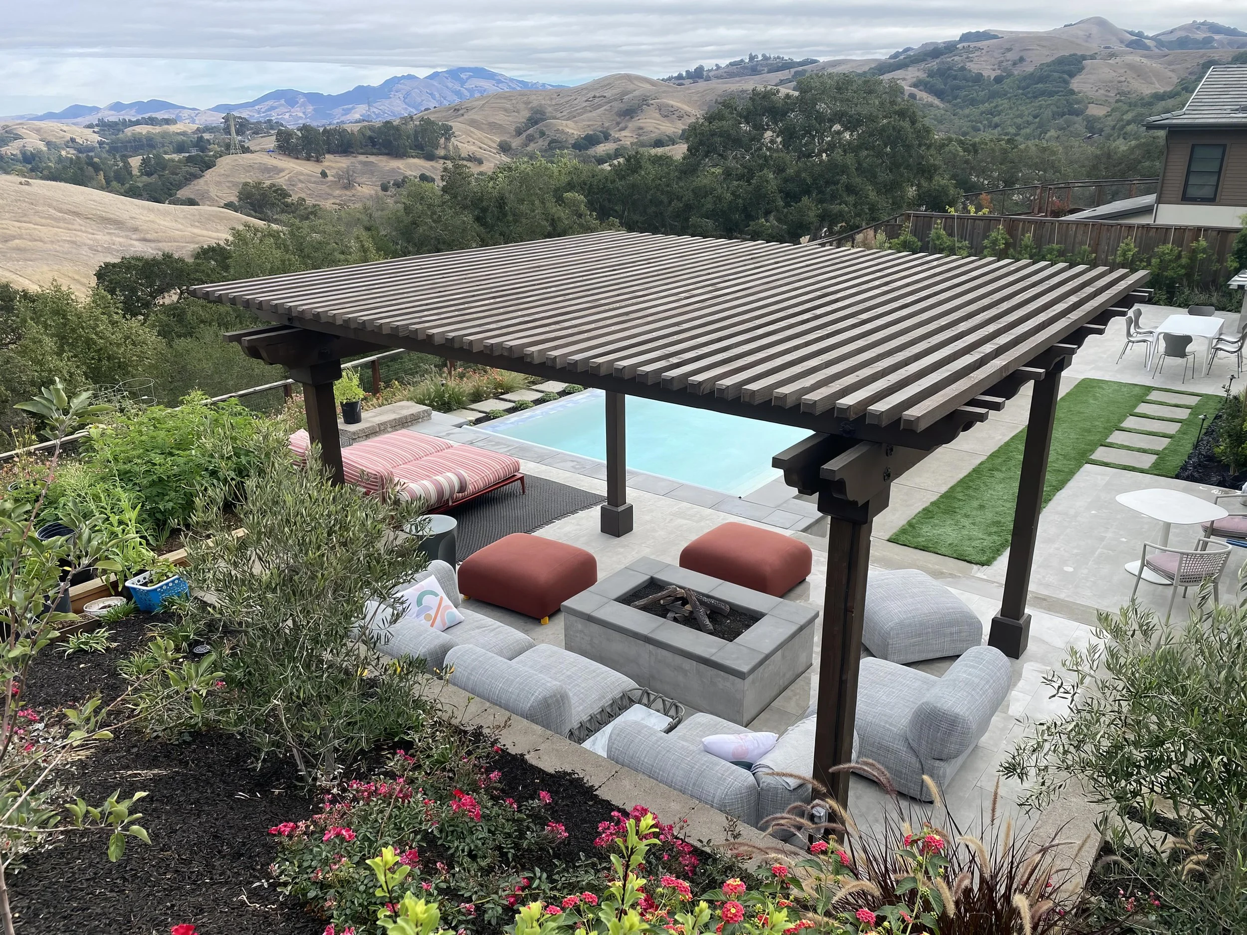 Outdoor backyard with a swimming pool, covered seating area with couches and ottomans, fire pit, patio furniture, and scenic mountains in the background.