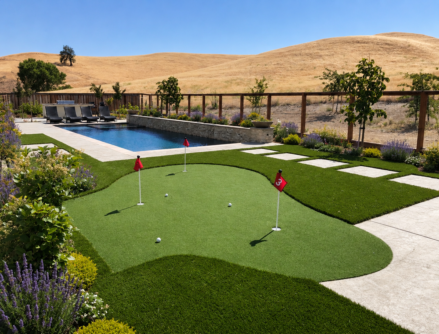 Backyard with a pool, a putting green with golf balls and flags, lounge chairs, and a garden with various plants and flowers, with dry hills and a tree in the background under a clear blue sky.
