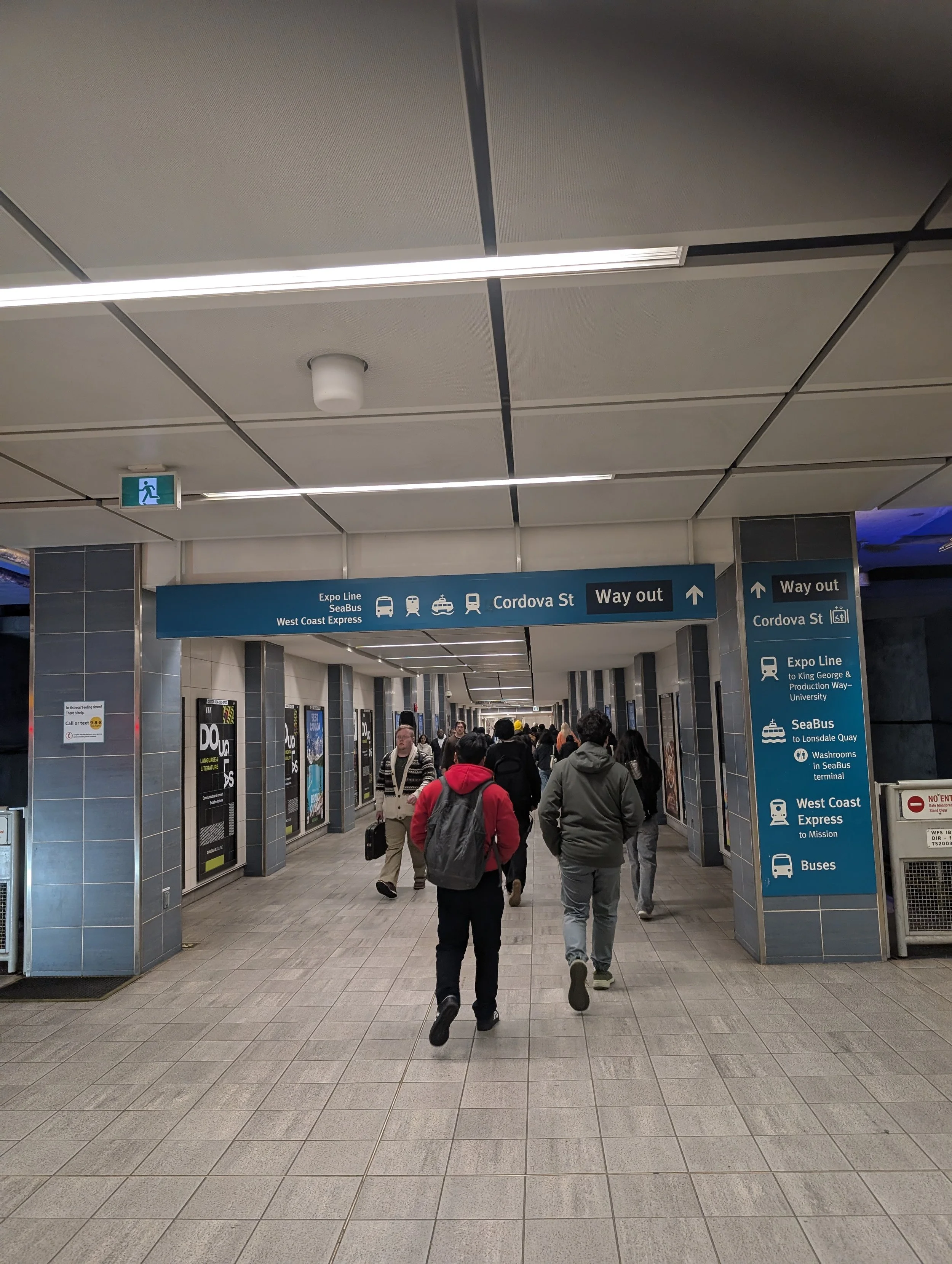 People walking through a corridor at a transportation terminal with signs indicating directions to various transit options including Expo Line, SeaBus, West Coast Express, buses, and Way out.