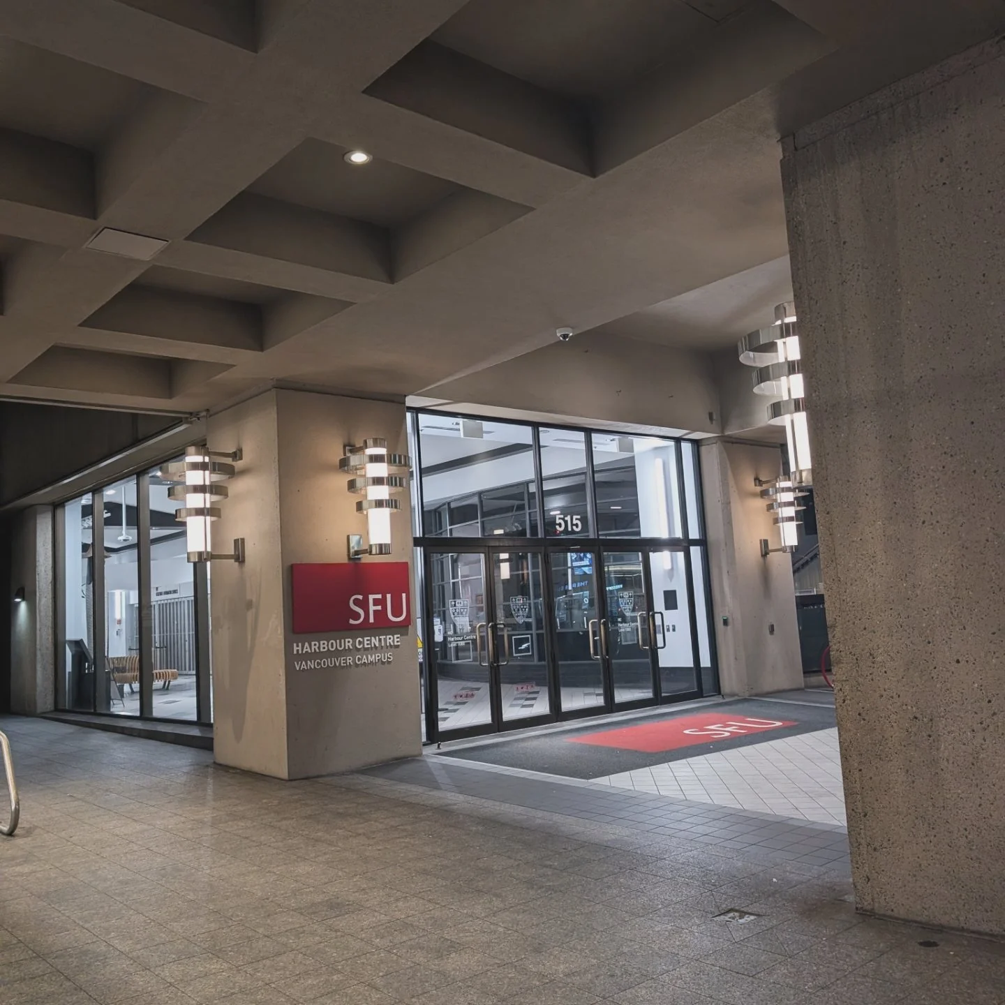 Entrance to SFU Harbour Centre Vancouver Campus with glass doors, a red SFU sign, and modern wall sconces, indoors in a gray concrete building.