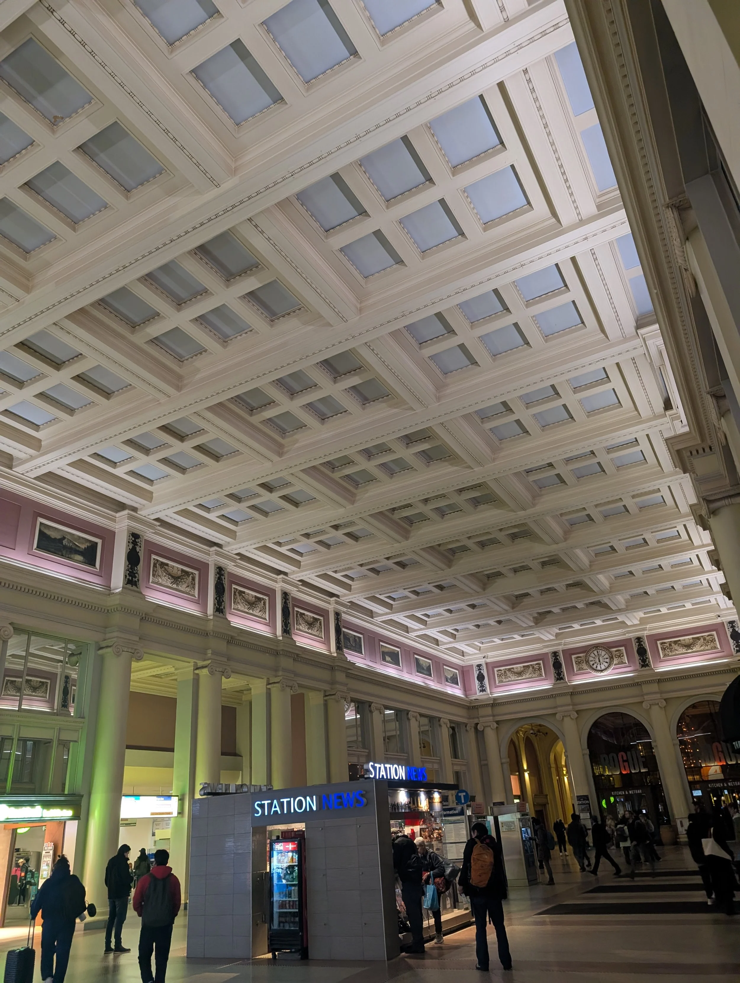 Interior of a grand train station with a high coffered ceiling, illuminated walls with moldings, and a newsstand labeled 'STATION NEWS'. People are walking and standing around.