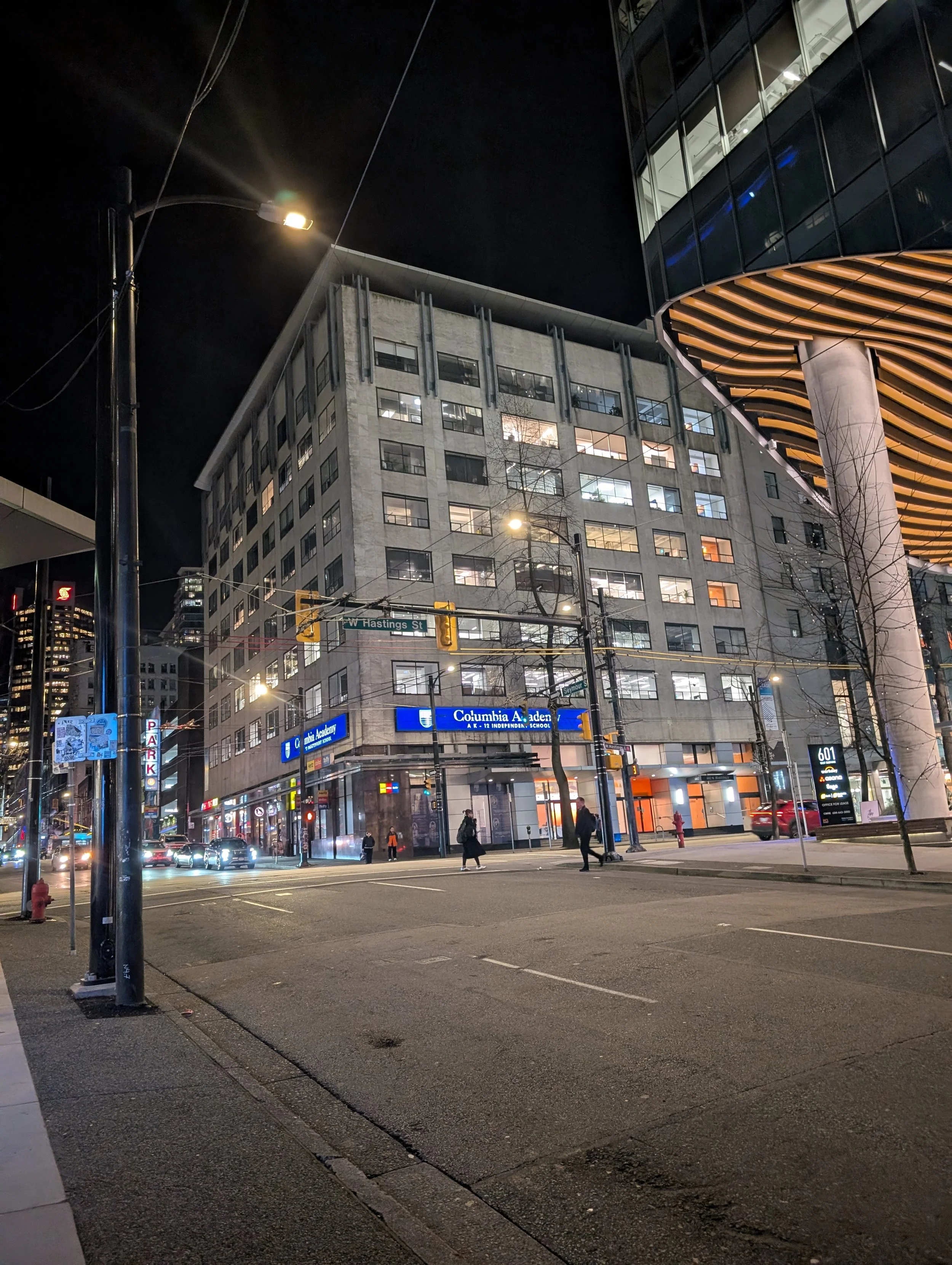 Nighttime city street scene with a multi-story building, illuminated signs, and people walking across the street.