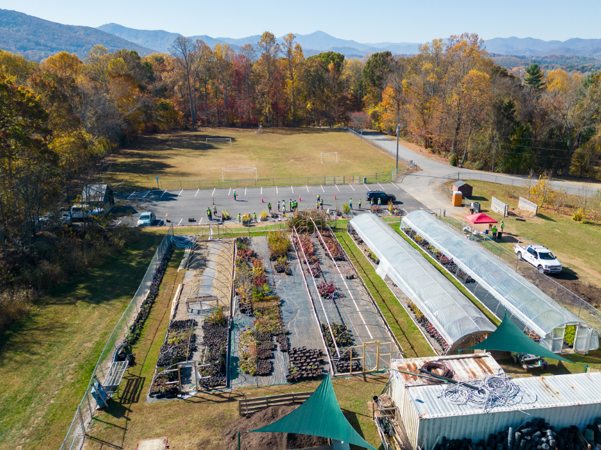 Aerial view of potted tree nursery showing two hoop houses and three long, rectangular concrete pads, all containing potted trees of varying species. Fall tree foliage and the Blue Ridge Mountains can be seen in the top third.