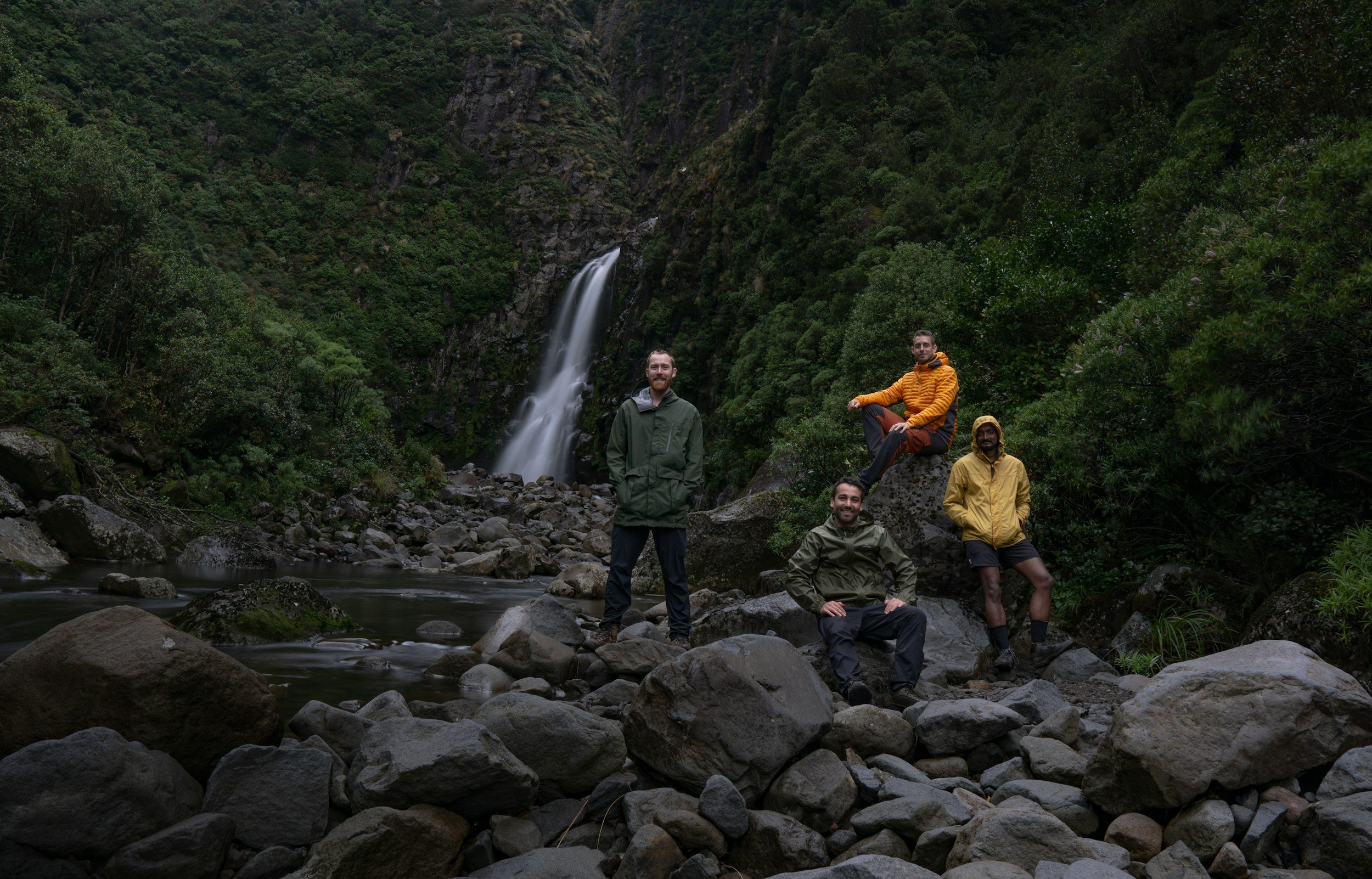 Four people posing in front of a waterfall in a lush green forest, wearing outdoor jackets and standing on rocks.