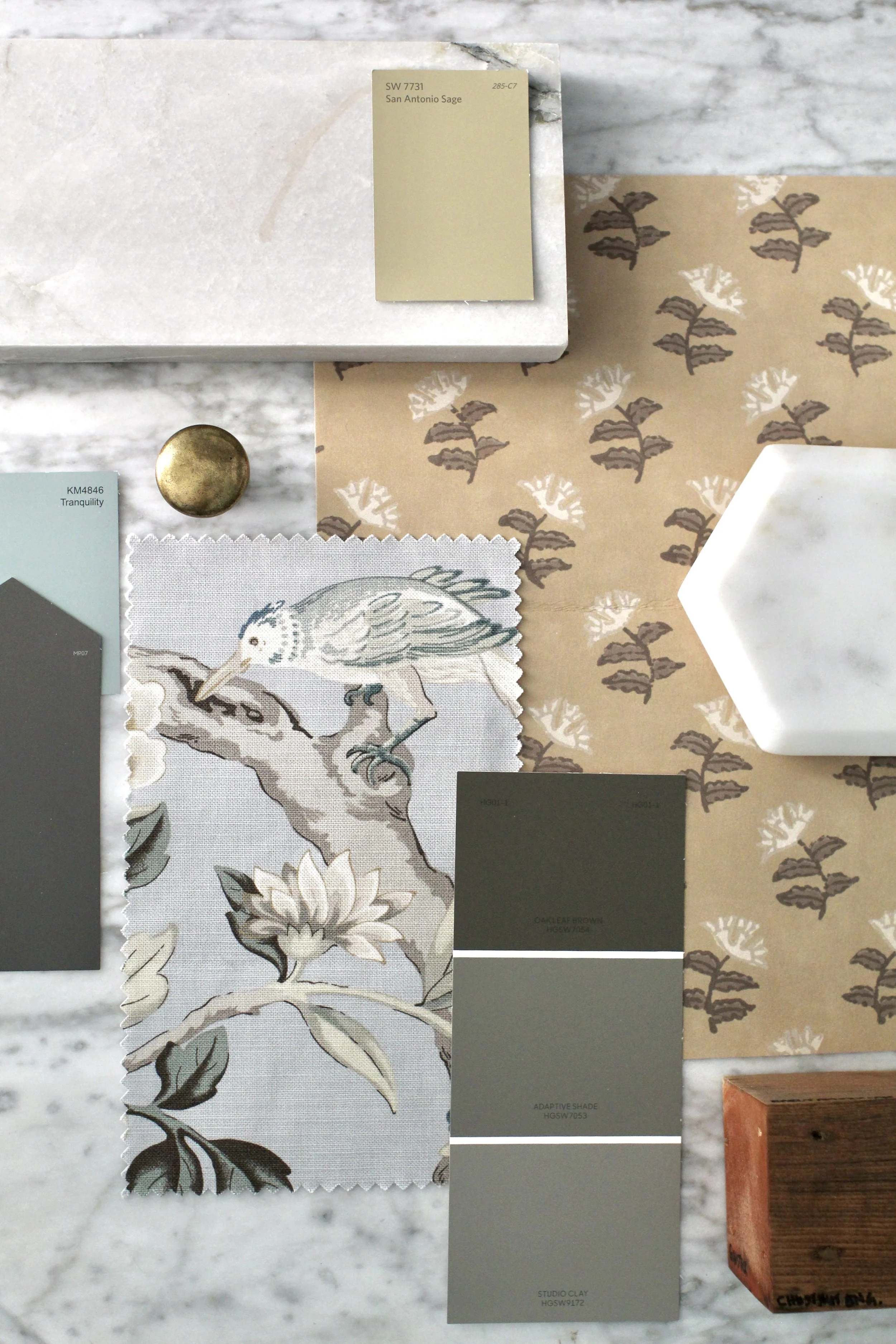 A collection of design samples, including a tile with a bird and floral motif, paint swatches in shades of gray and green, and wallpaper or fabric with a leaf pattern, arranged on a marble surface.