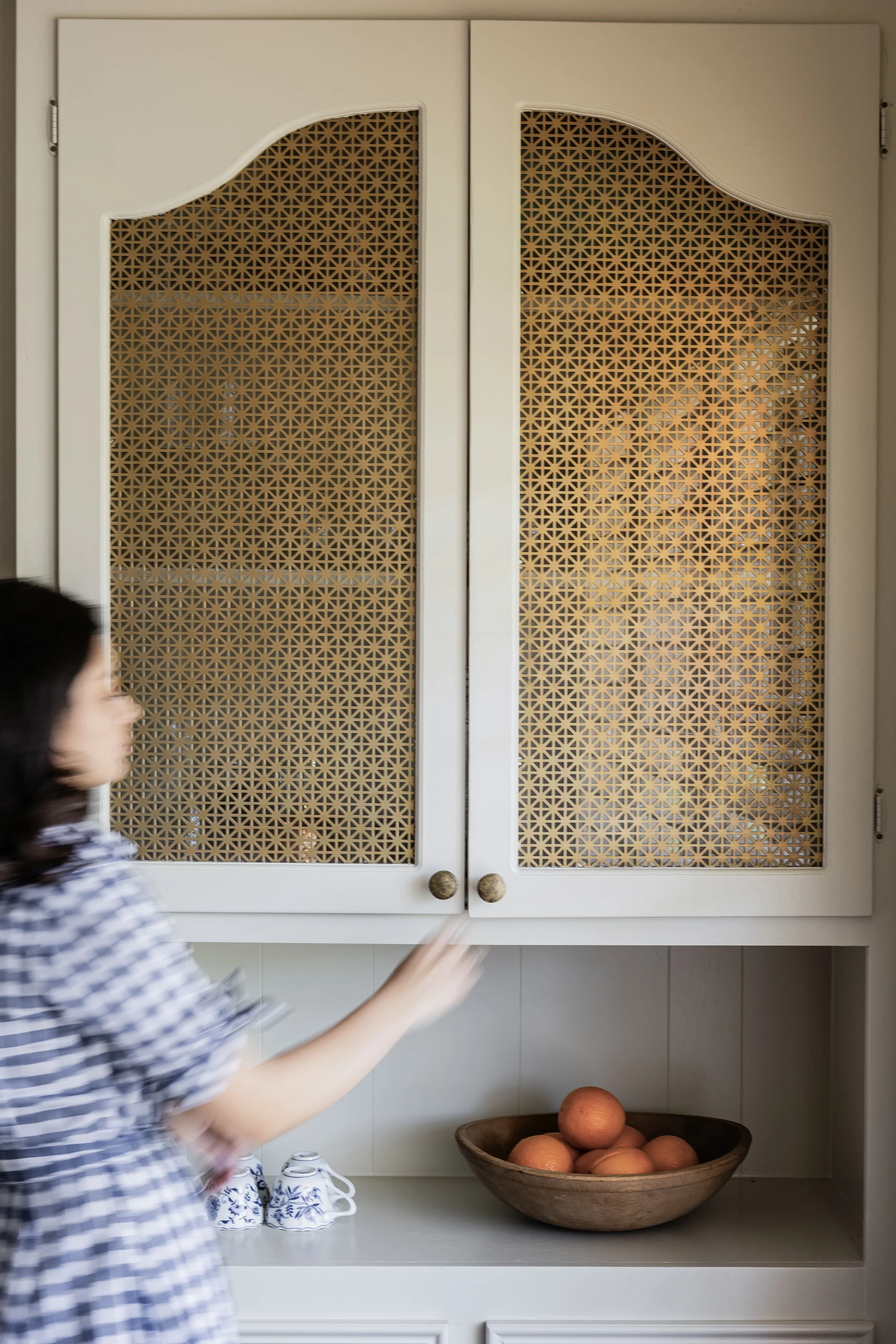 A kitchen cabinet with a decorative lattice design on the doors, above a countertop. A woman in a checkered shirt is reaching for an object. On the counter, there are three cups with blue and white patterns and a bowl of large round brown objects, possibly oranges. In the background, part of a person and a wall with kitchen items are visible.