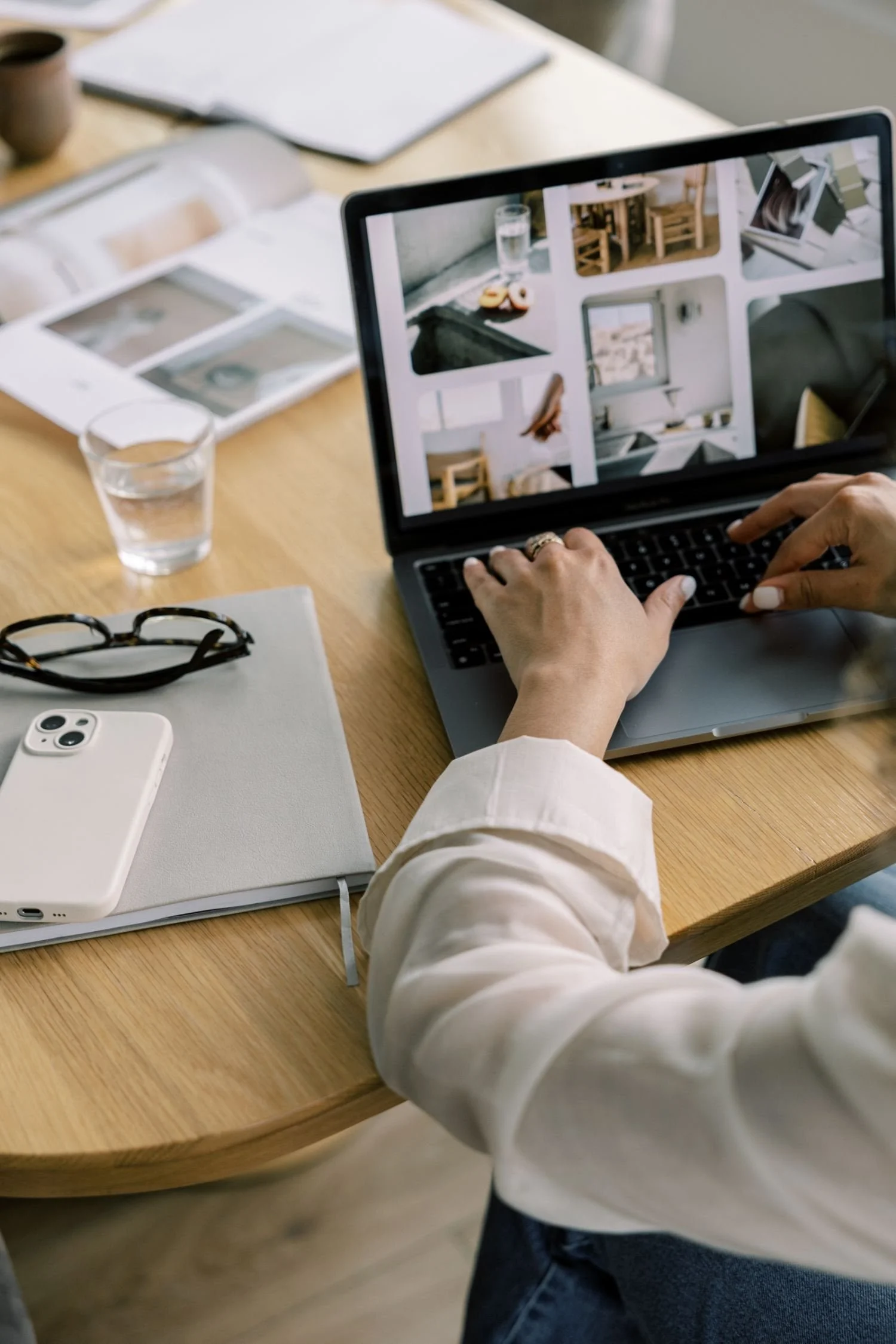 A person in a white long-sleeve shirt is working on a laptop at a light wood desk, arranging or viewing multiple images of interior spaces on the screen. The desk has a white notebook, a pair of glasses, a white smartphone, a glass of water, and some printed photographs or documents. There is also a cup and a notebook in the background.