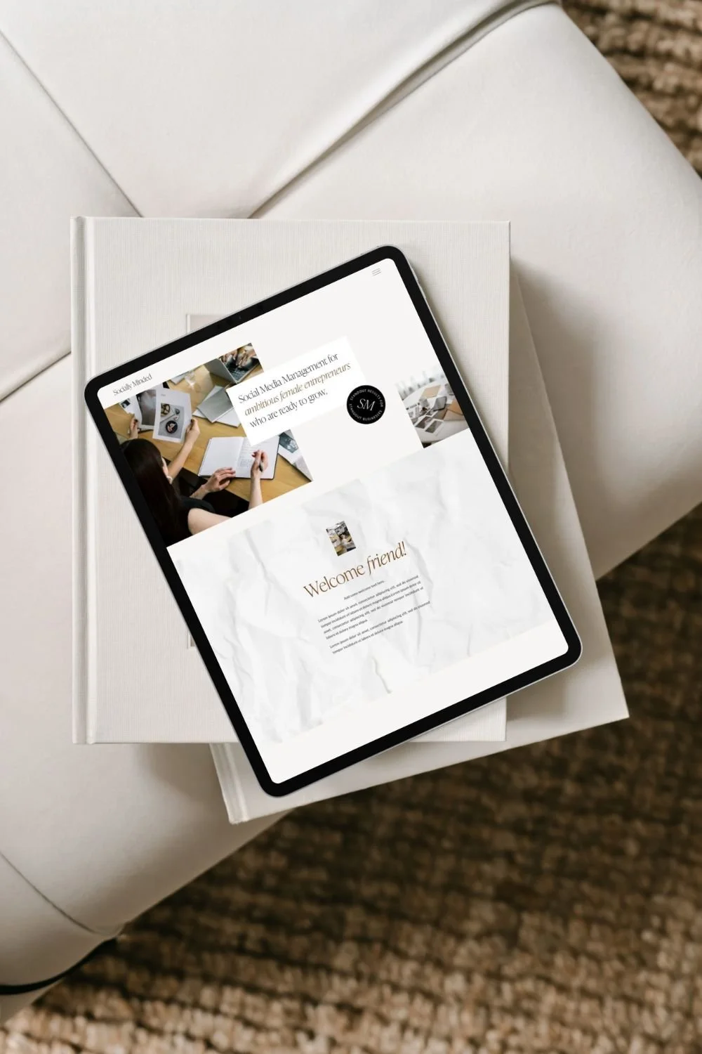 A tablet displaying a website with images of women working at desks, placed on top of a closed white book on a white table with a textured brown carpet below.