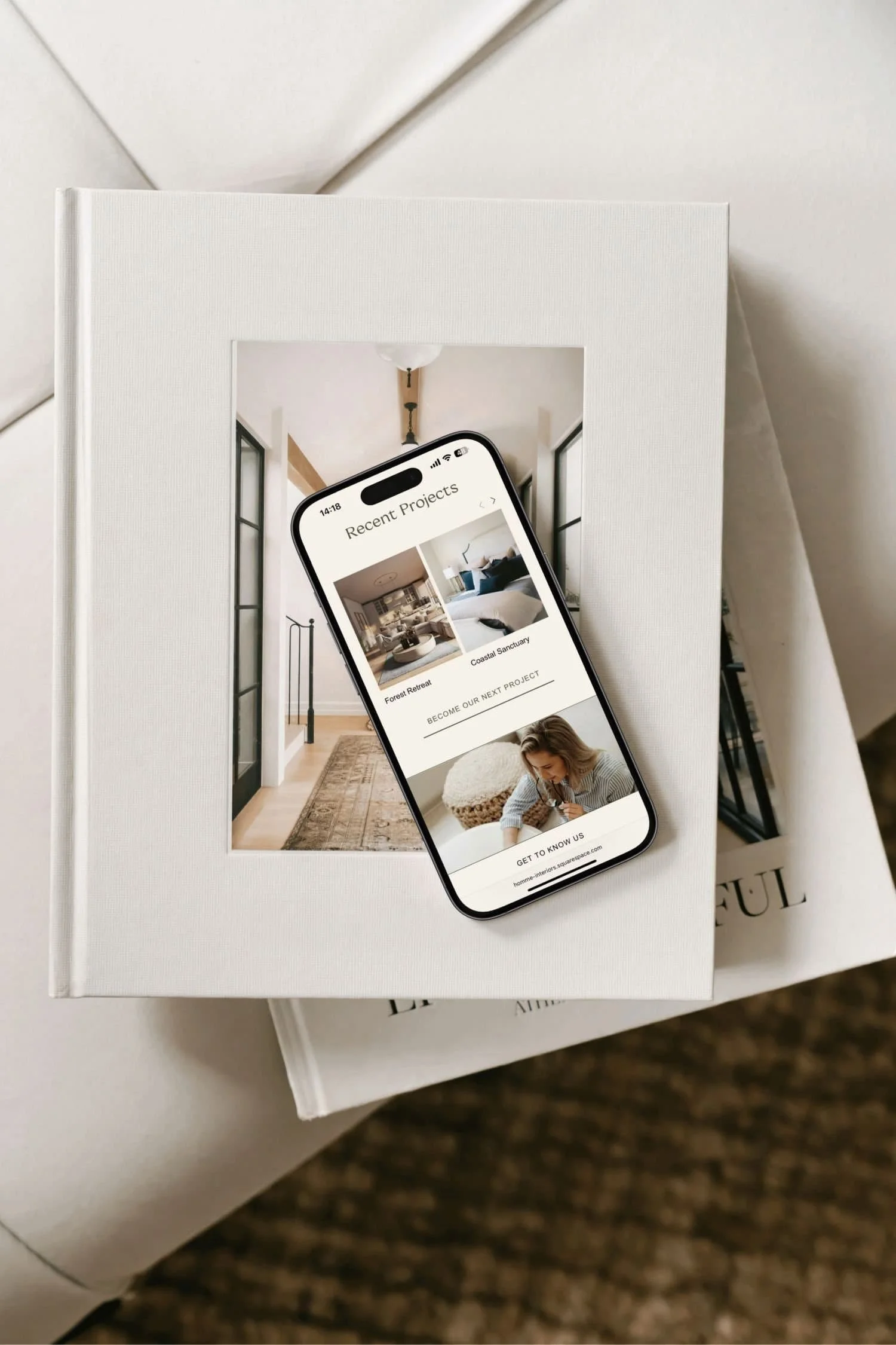 A white photo book with a mirror on the cover, and a smartphone resting on top displaying a website for interior design projects. The website shows images of modern home interiors with text overlaid.