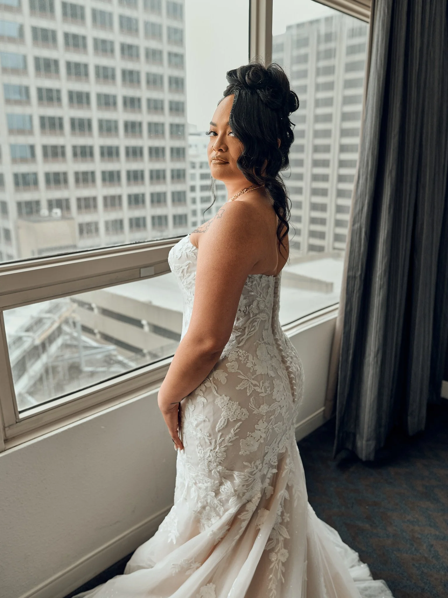 A woman in a white and floral lace wedding dress standing by a large window in a high-rise building.