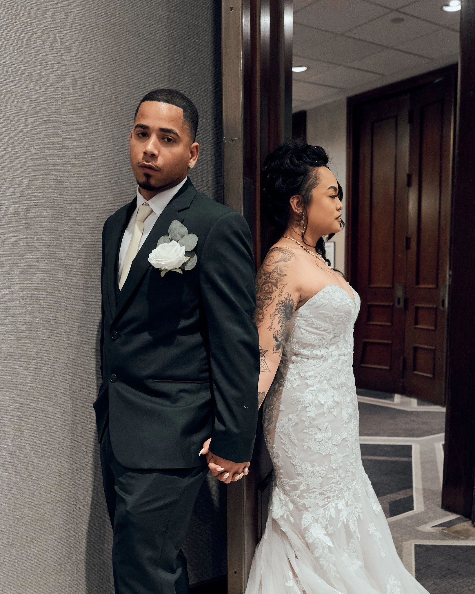 A couple in wedding attire, standing back to back, holding hands, in an indoor setting with wood panel doors in the background.
