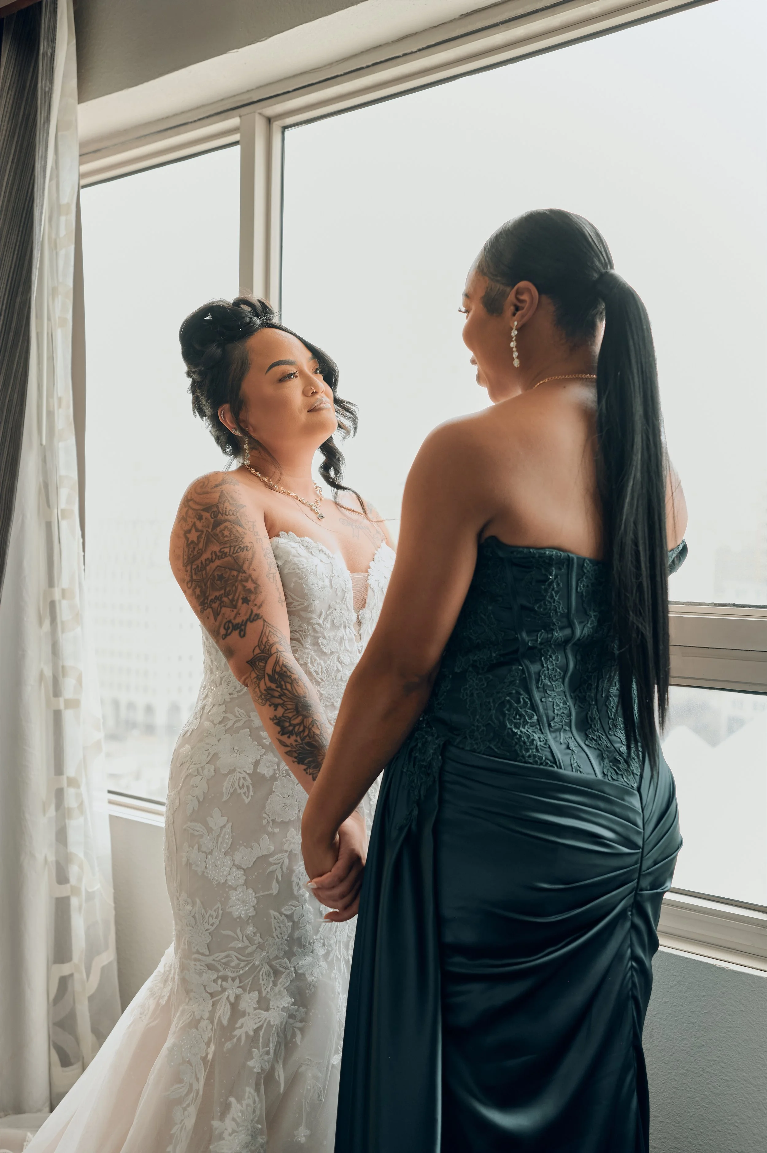 Two women holding hands and gazing at each other by a window, one wearing a white wedding dress with floral details and tattoos, and the other wearing a strapless dark gown with long black hair.