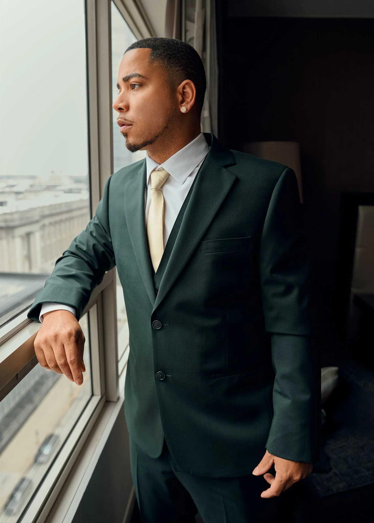 A young man in a dark suit, light dress shirt, and light tie looking out the window in a high-rise building.