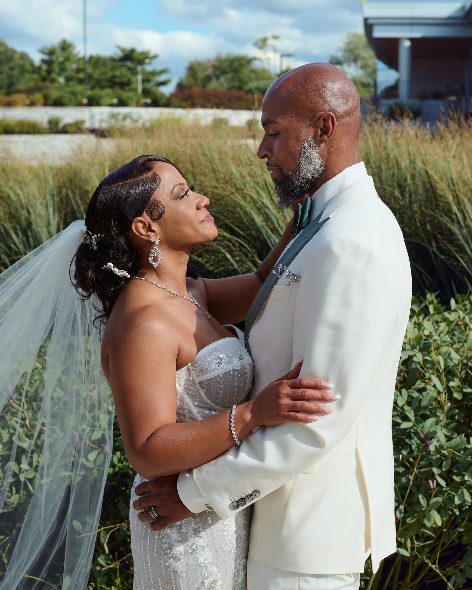 A bride and groom in wedding attire embrace outdoors, with trees and shrubbery in the background.