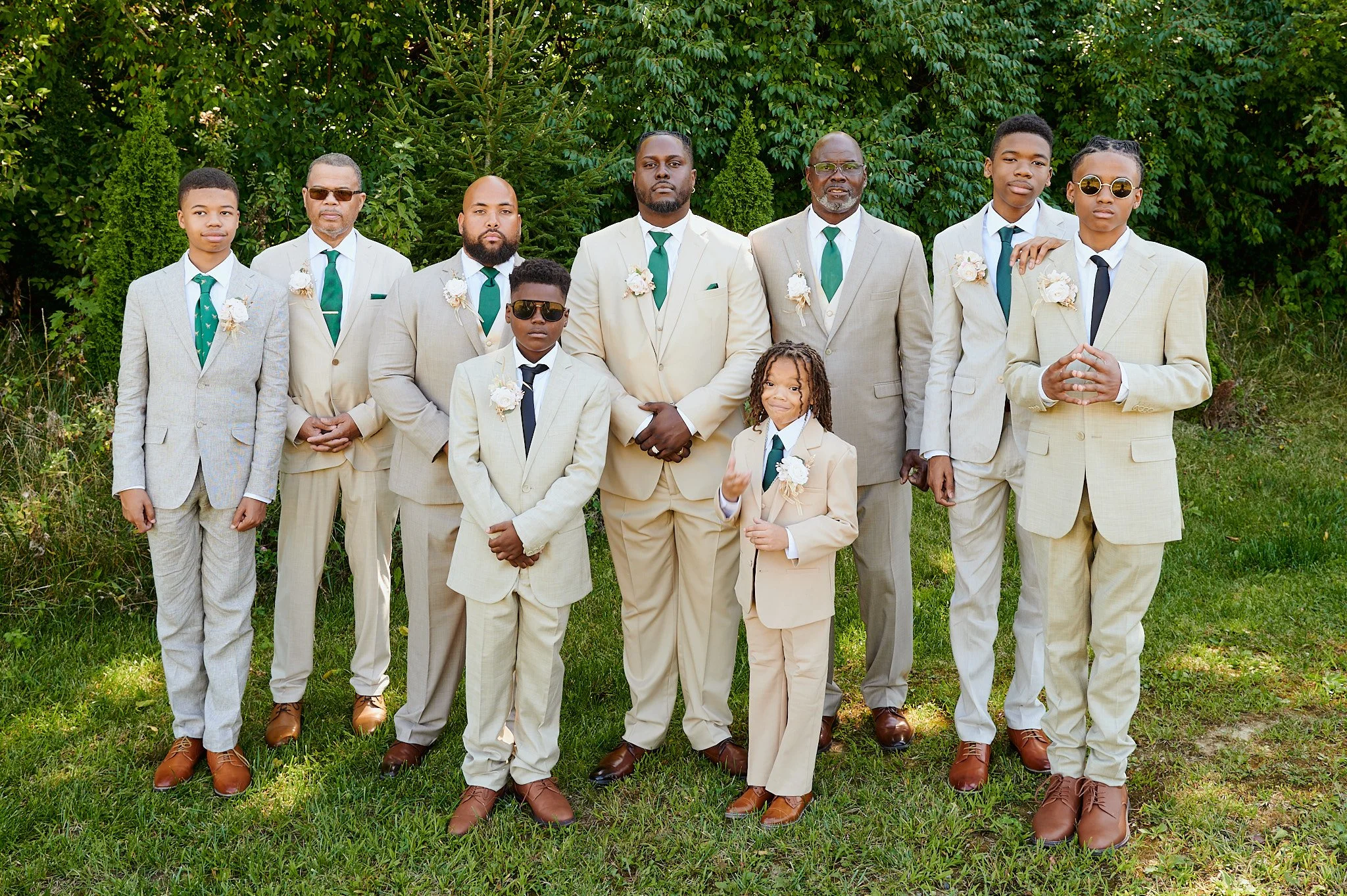 A group of ten men and boys dressed in light-colored suits with green ties, standing outdoors on grass with green trees in the background. Some are wearing sunglasses, and they have boutonnieres.