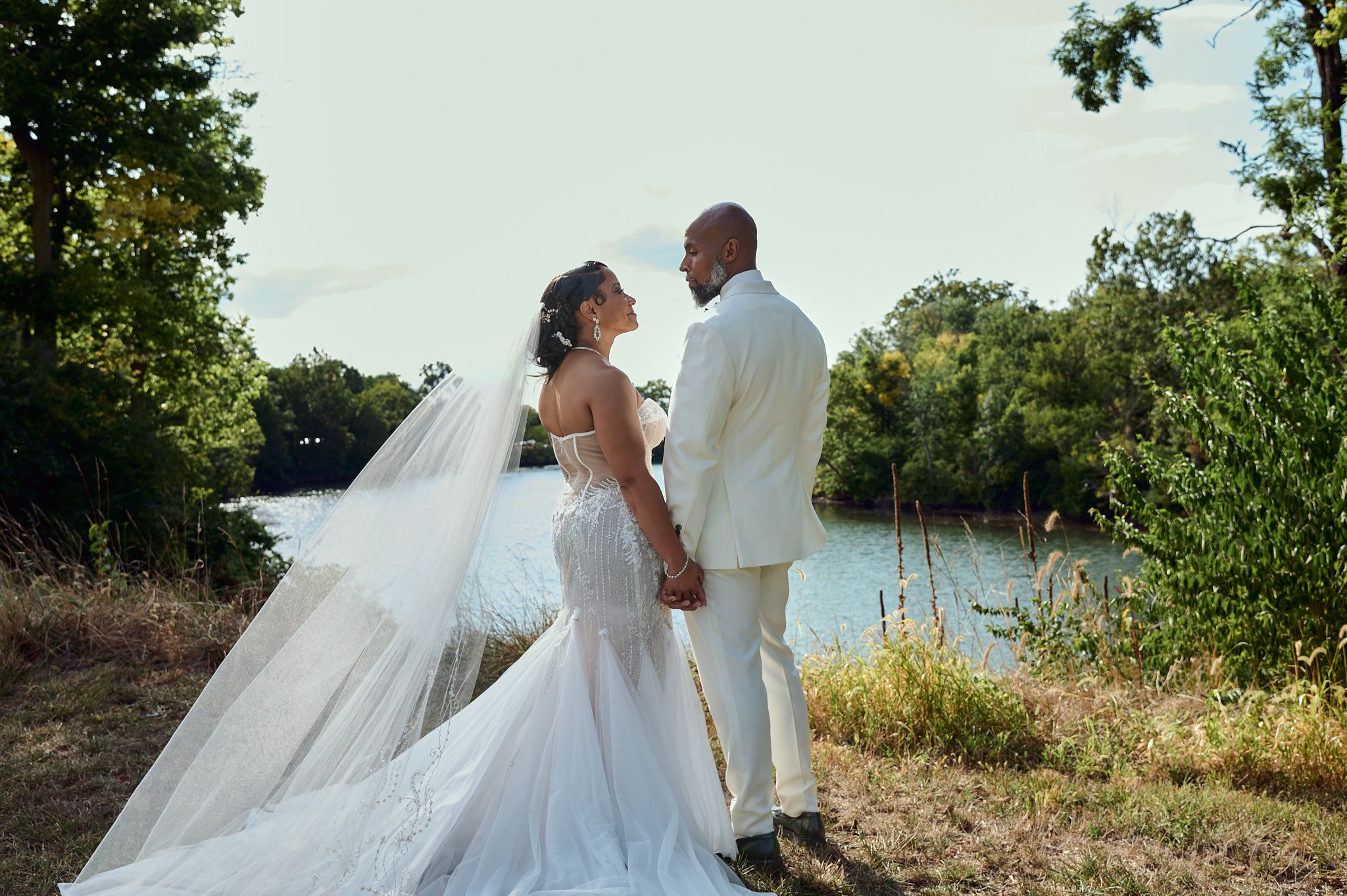 A bride and groom holding hands by a river, dressed in wedding attire, surrounded by greenery on a sunny day.