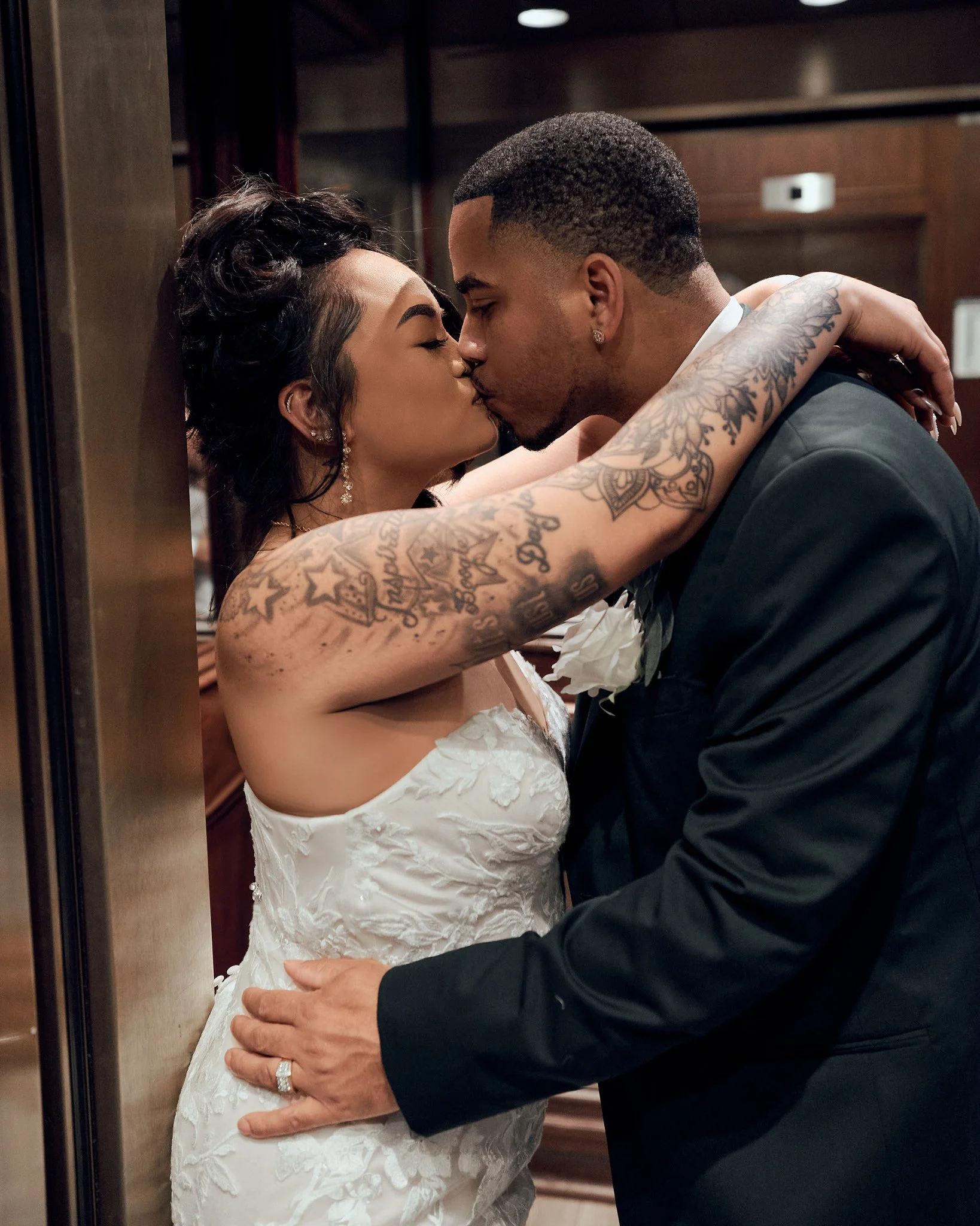 A newly married couple sharing a kiss in an elevator, the woman in a white wedding dress with tattoos on her arm, the man in a black suit with a wedding band.