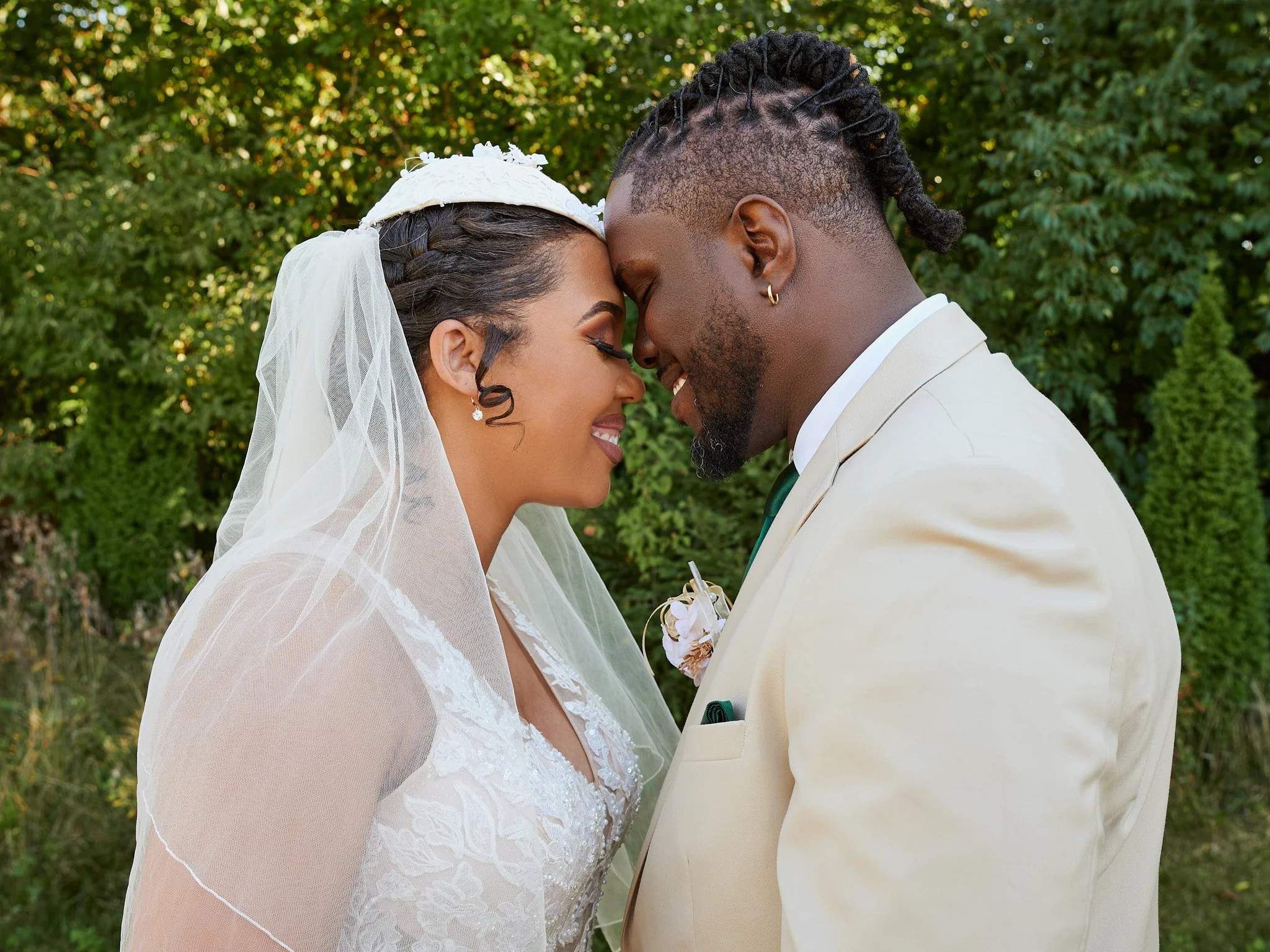 Bride and groom with foreheads touching, smiling, outdoors with green trees in background.