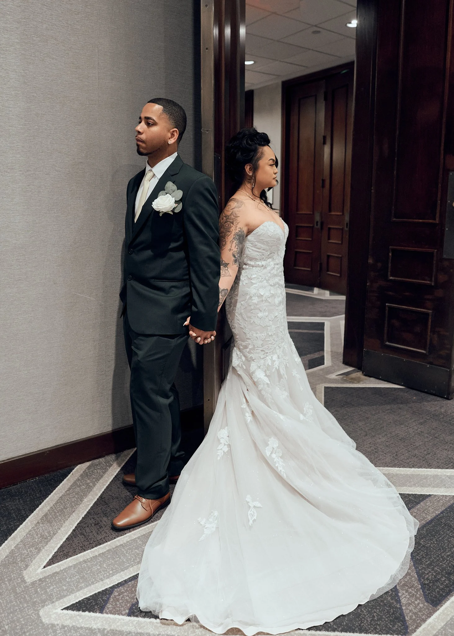A bride and groom with their backs against each other, holding hands, in a hotel hallway. The bride is wearing a strapless white lace wedding gown with a long train, and the groom is dressed in a black suit with a white shirt and tie.