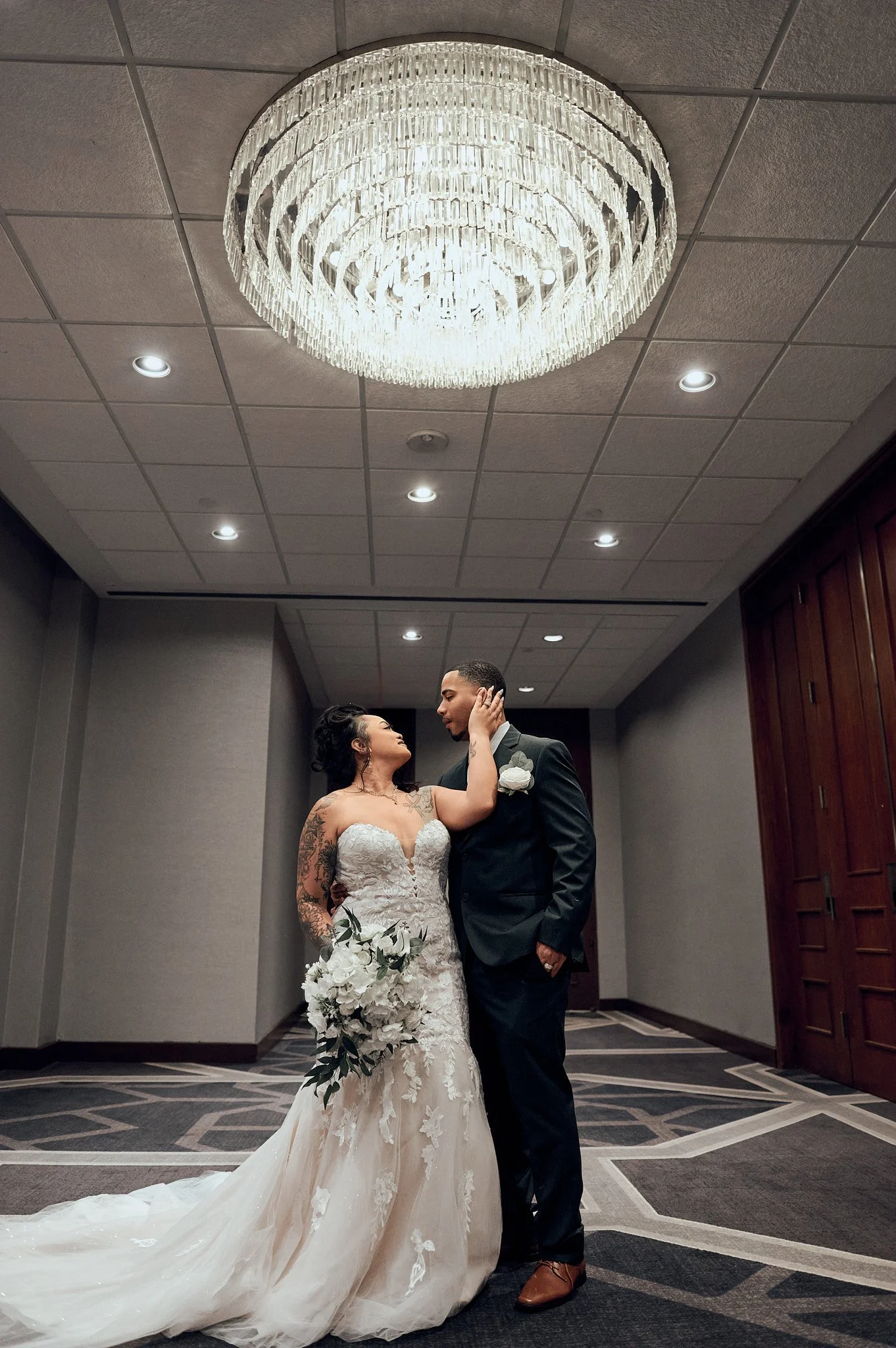 A bride and groom standing close on their wedding day, looking into each other's eyes, in an elegant indoor setting illuminated by a large crystal chandelier.