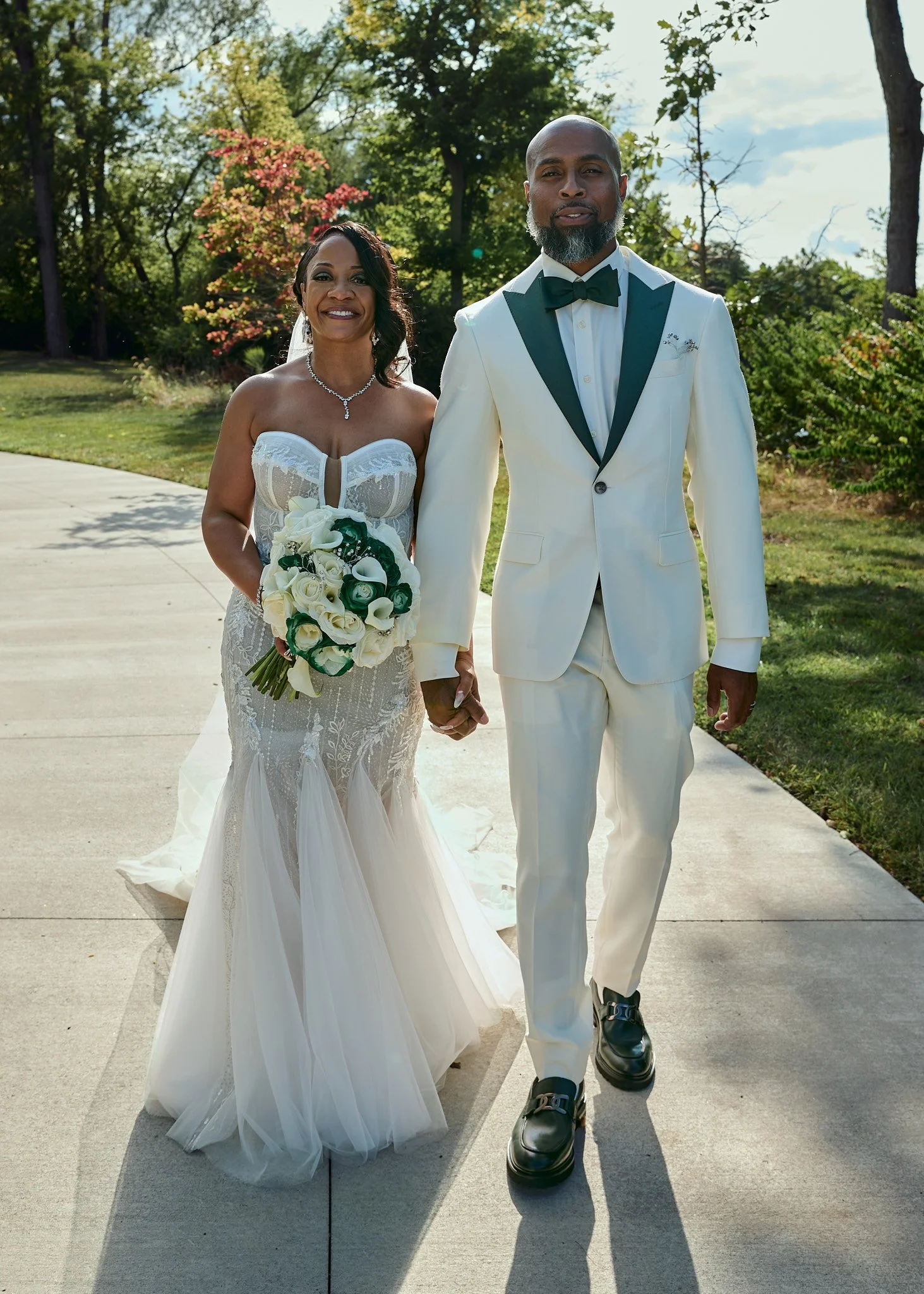 A bride and groom walking hand in hand outdoors on a sunny day, dressed in wedding attire. The bride wears a strapless white lace wedding gown and holds a bouquet of white roses and greenery. The groom wears a white tuxedo with a black collar, black 