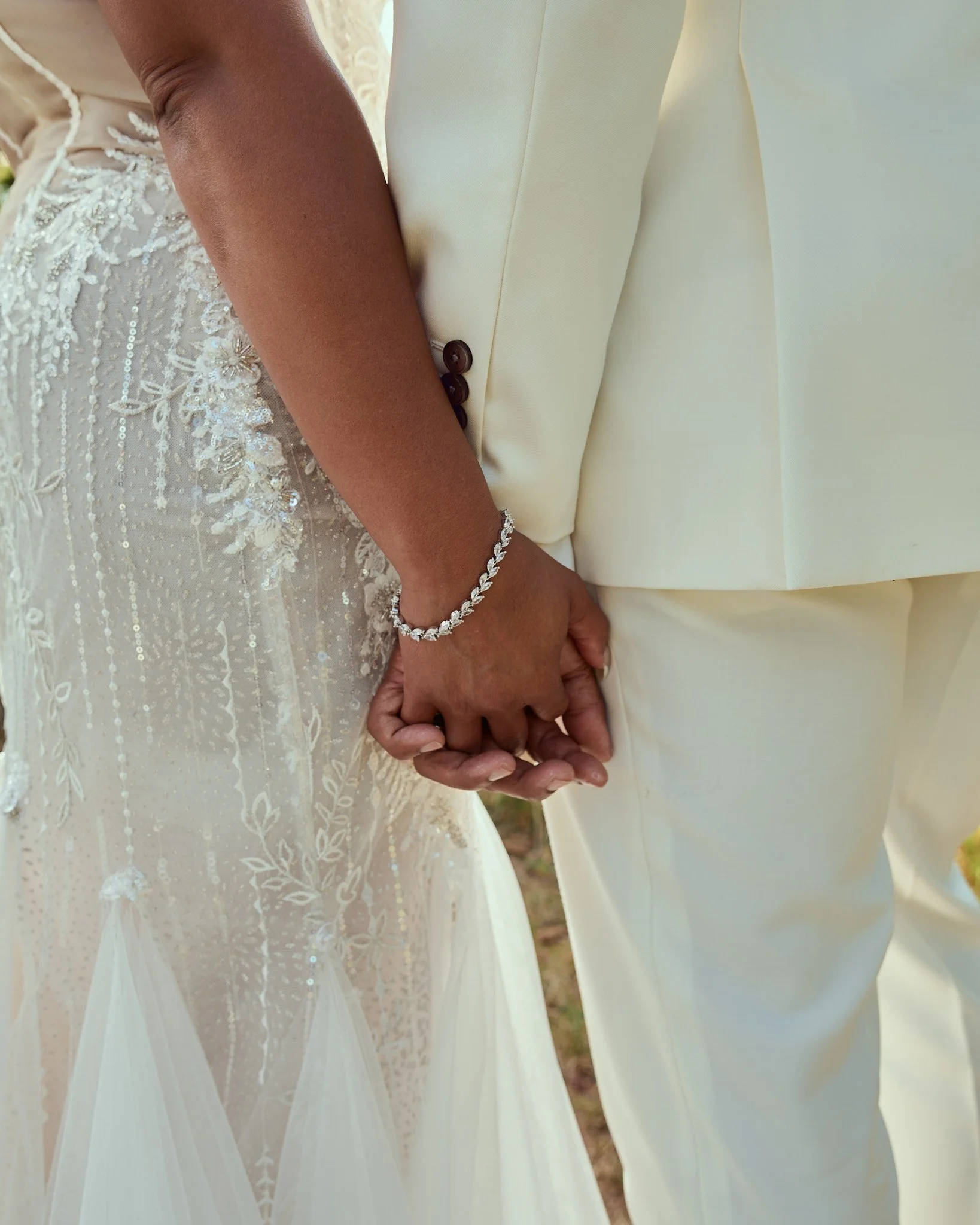 Close-up of a bride and groom holding hands, with the bride wearing a lace wedding dress and shimmering embellishments, and the groom in a cream suit. The bride wears a diamond bracelet and the groom's shirt has dark buttons.